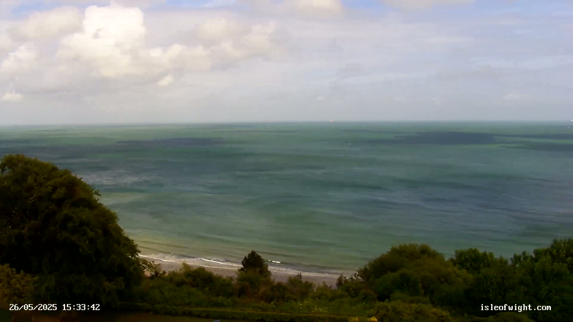 A wide expanse of greenish-blue ocean under a partly cloudy sky stretches to the horizon, with a sandy beach and lush green vegetation visible in the foreground.