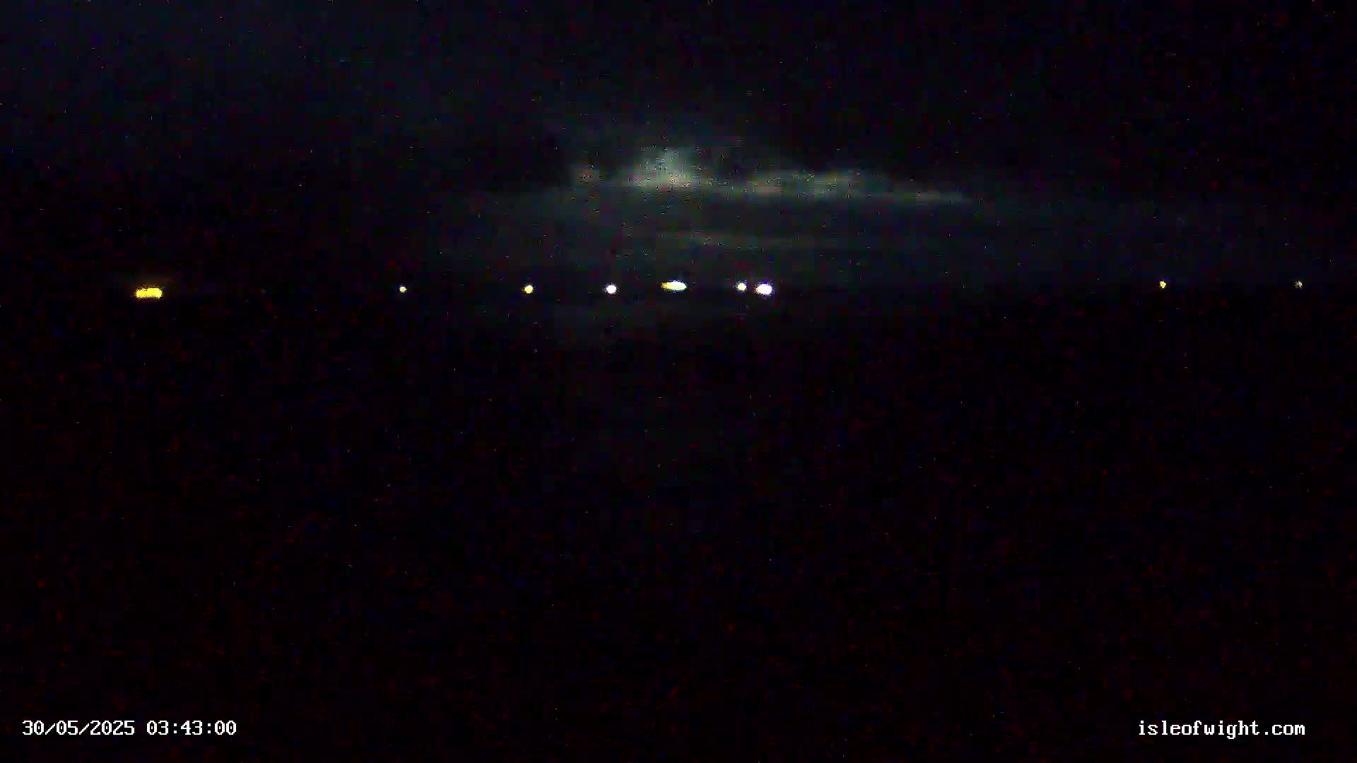 A nighttime, dark scene shows a low horizon line with several distant, faint lights under a mostly cloudy sky.