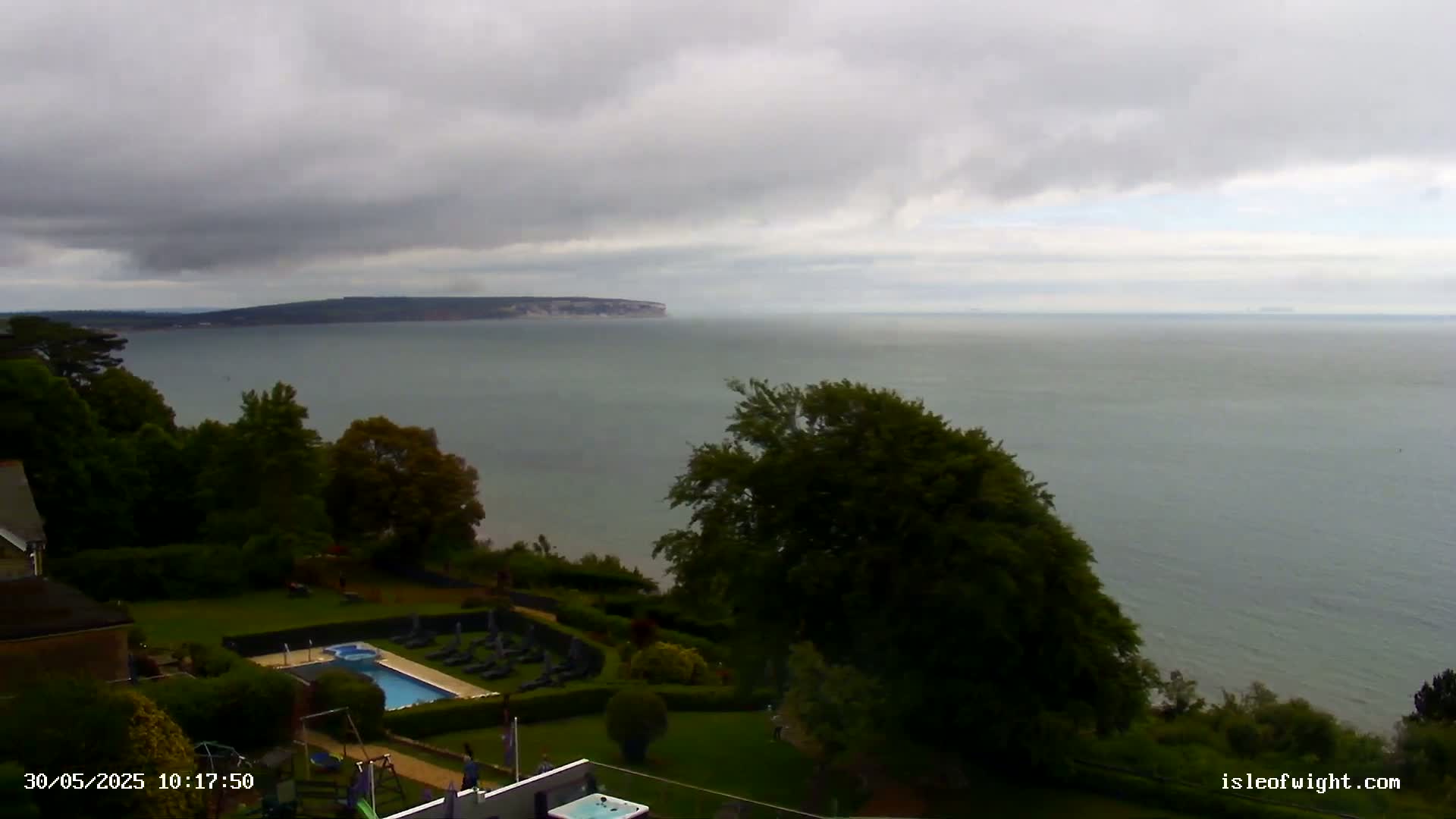 An overcast day reveals a coastal view of a calm sea, a swimming pool and surrounding lawn, and distant land under a cloudy sky.