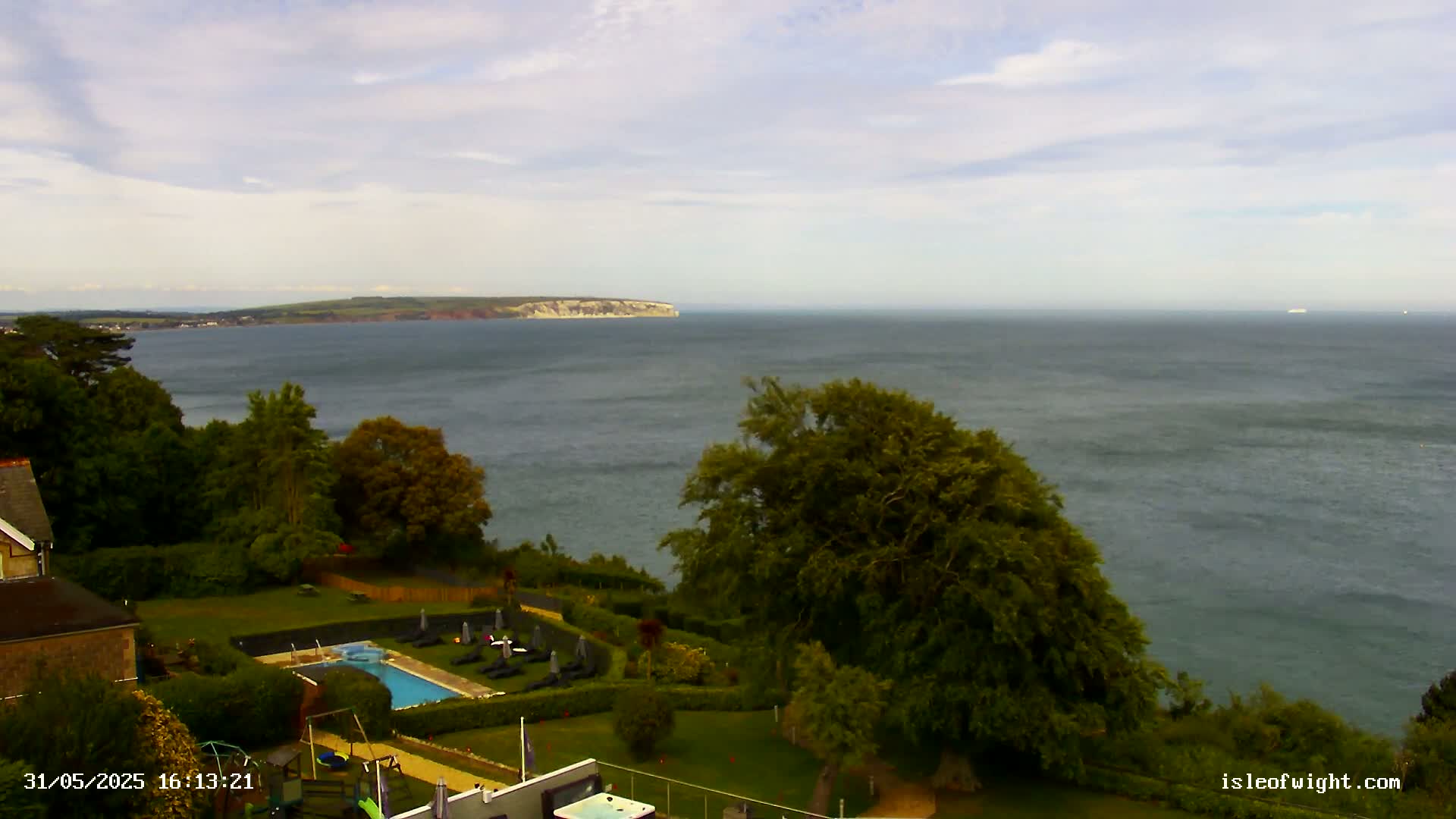 A coastal view shows a swimming pool and lawn area next to a cliff overlooking a calm sea and a distant landmass under a partly cloudy sky.