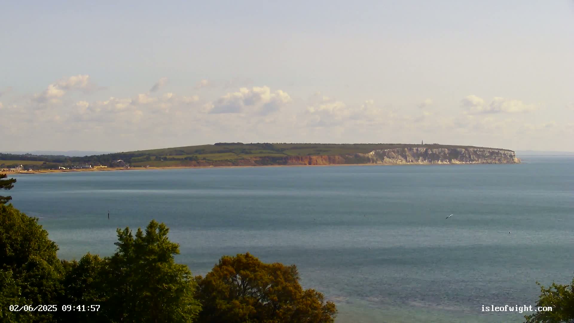A calm, partly cloudy day reveals a coastal scene with a body of water separating a tree-lined foreground from a distant shoreline featuring rolling green hills and a prominent cliff face.