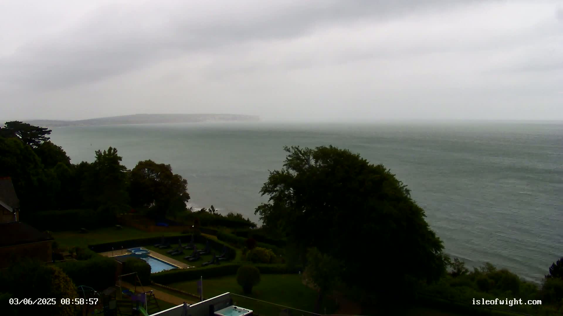 An overcast sky hangs over a calm sea viewed from a verdant hillside, where a swimming pool and other landscaping features are visible.