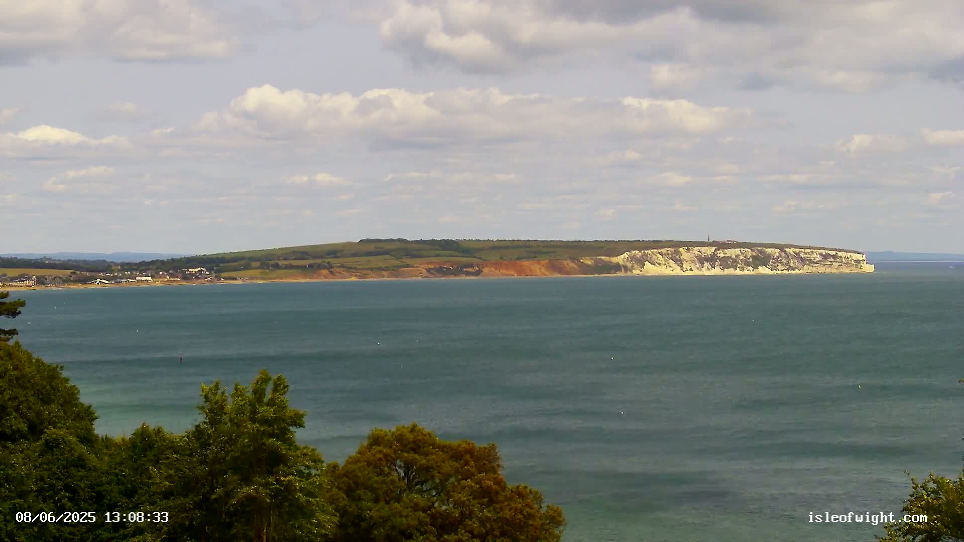 A partly cloudy day reveals a view of a calm body of water, with a distant coastline featuring rolling hills and a prominent cliff face.