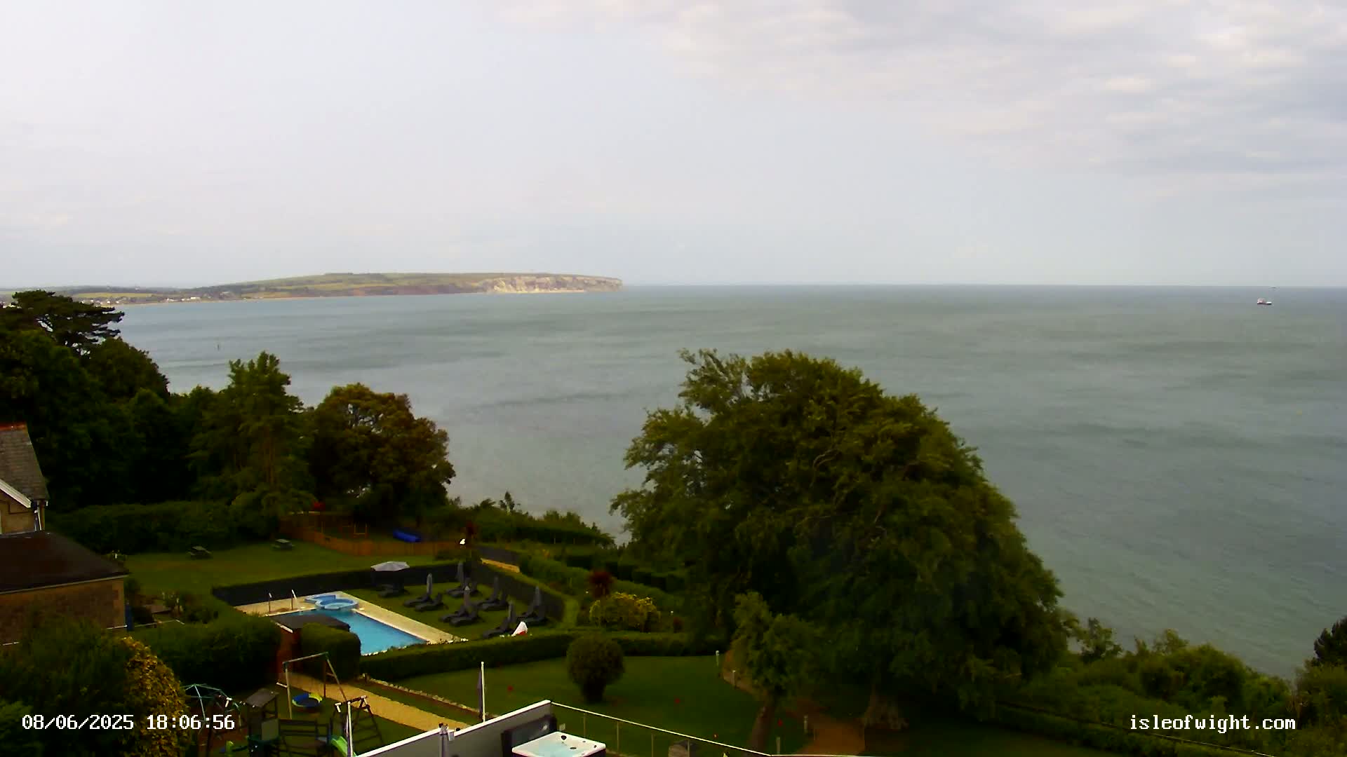 An overcast sky overlooks a tranquil sea with a distant landmass, while a swimming pool and surrounding greenery are visible in the foreground.