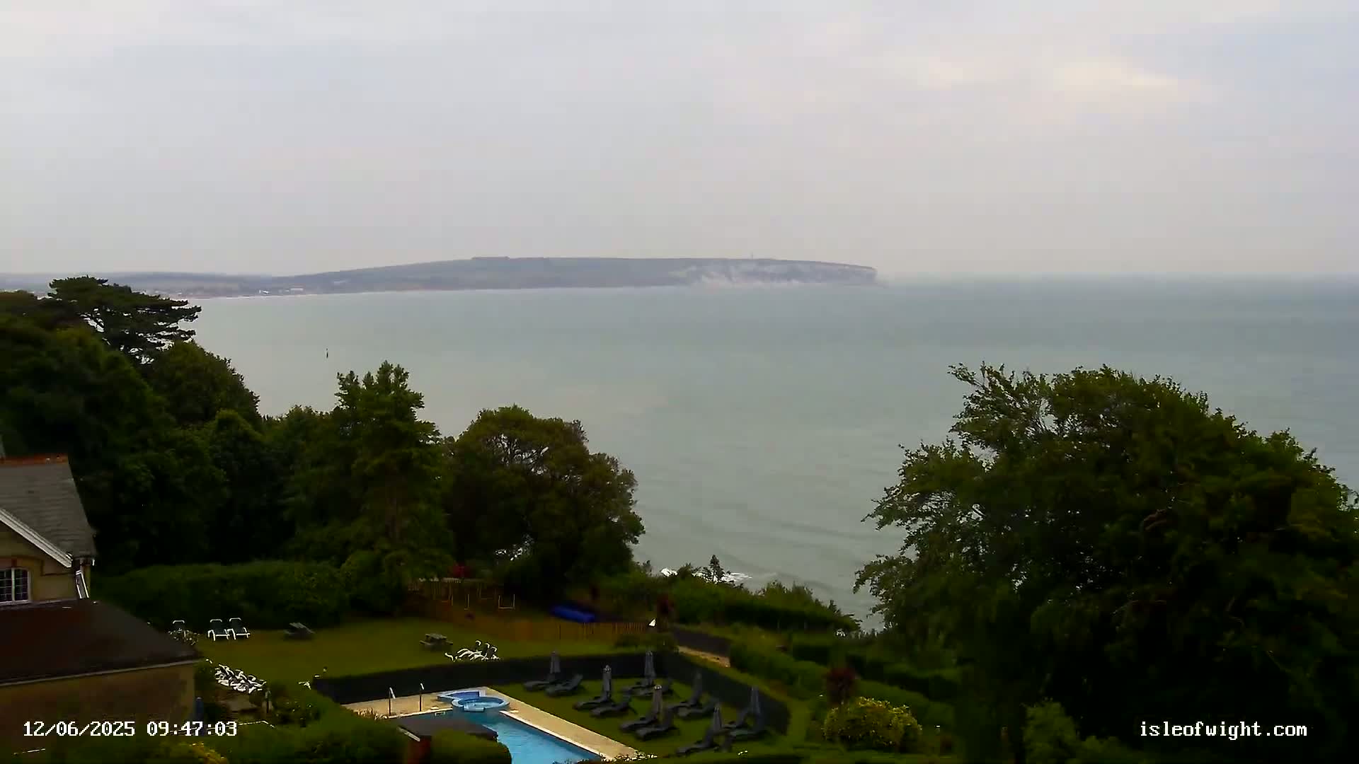 A hazy coastal scene shows a swimming pool and lawn next to a building overlooking a calm sea and distant land.