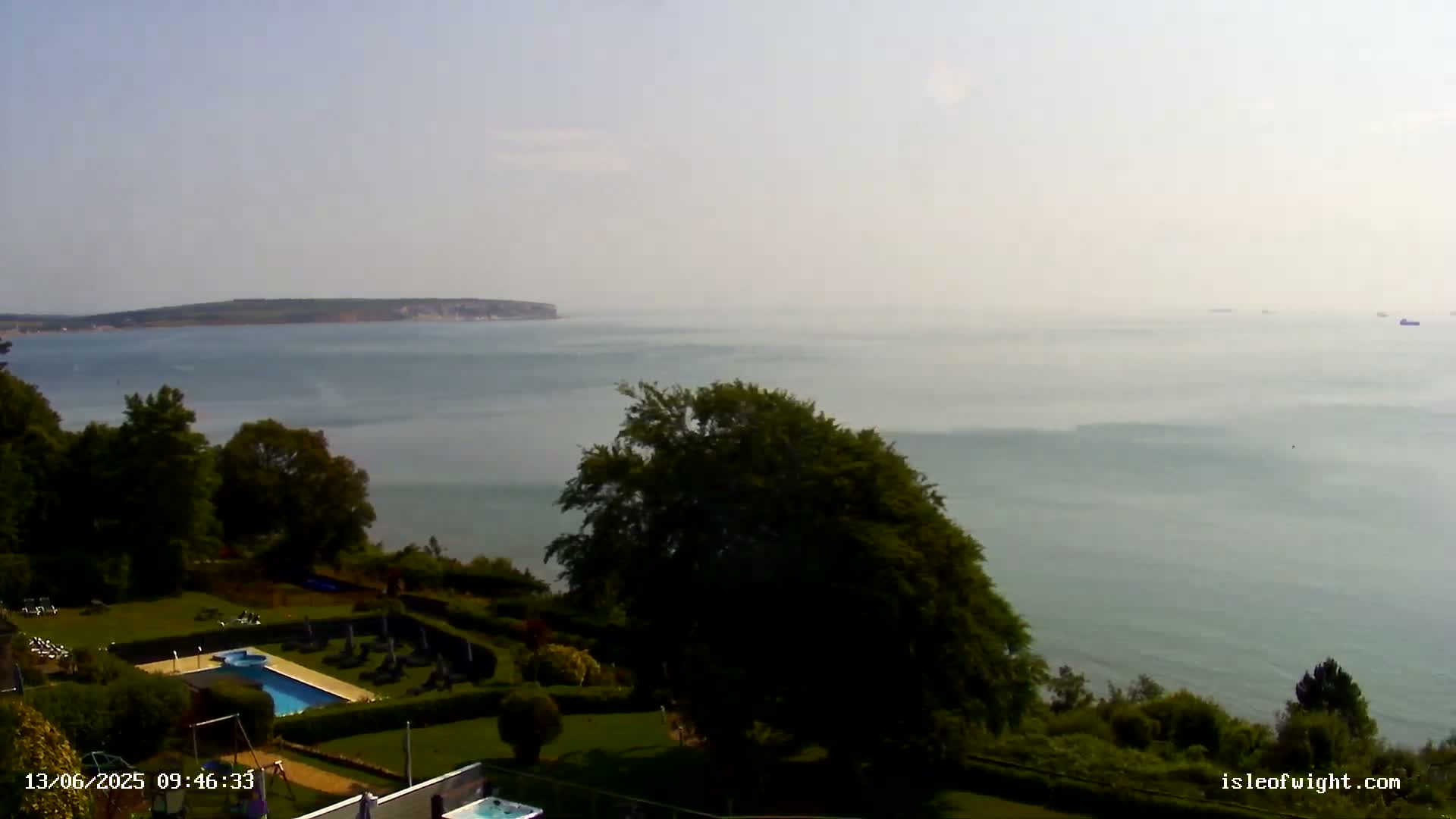 A hazy, sunny day reveals a coastal view of a swimming pool and manicured lawns overlooking a calm sea and distant landmass.