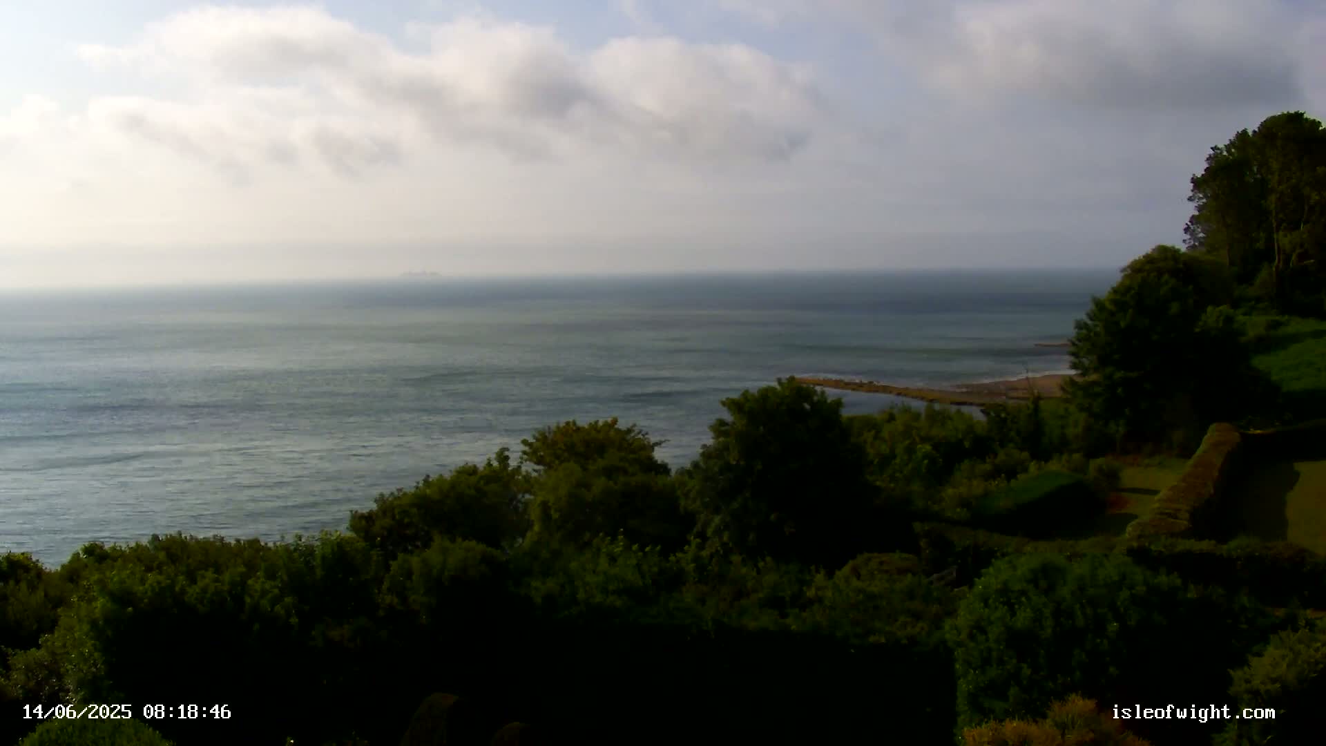 A partly cloudy day reveals a calm ocean view from a verdant hillside, with dark green trees and shrubs in the foreground.