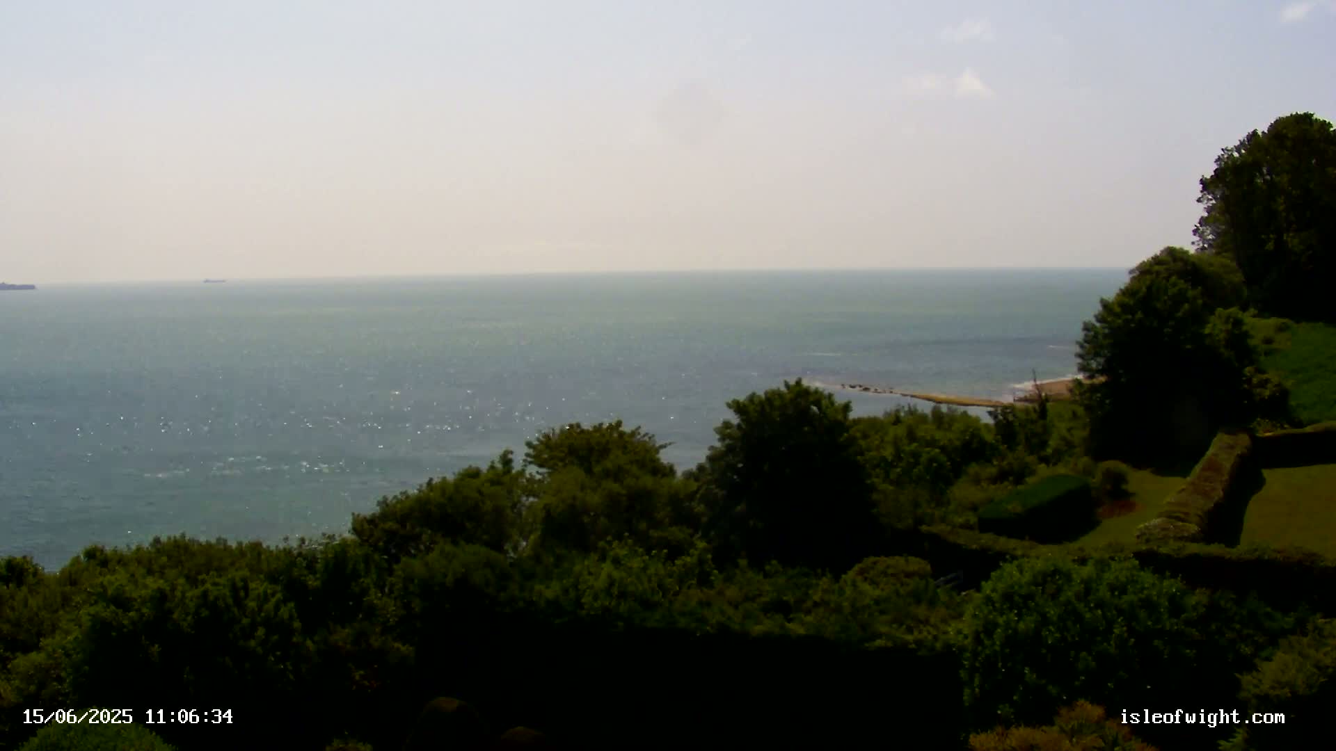 A calm, hazy ocean stretches to the horizon, viewed from a high vantage point overlooking lush green vegetation and manicured hedges.