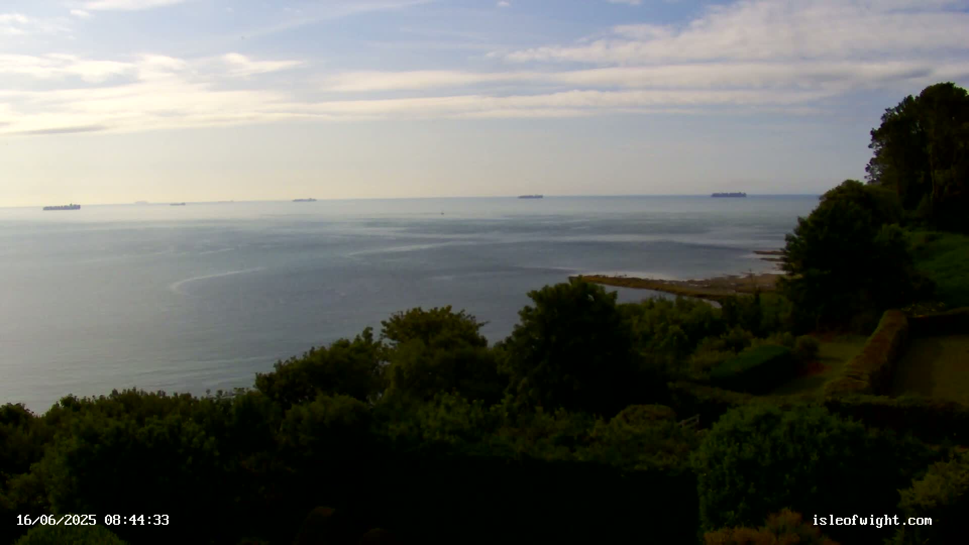 A calm ocean with several distant ships is viewed from a high vantage point overlooking a lush green landscape under a partly cloudy sky.