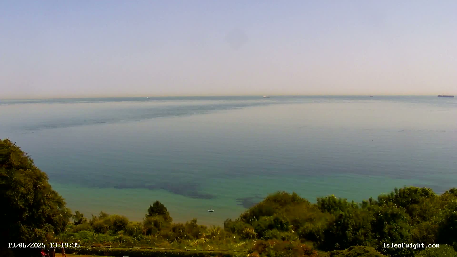 A hazy, sunny day overlooking a calm, teal-colored ocean with a few distant ships and green vegetation in the foreground.