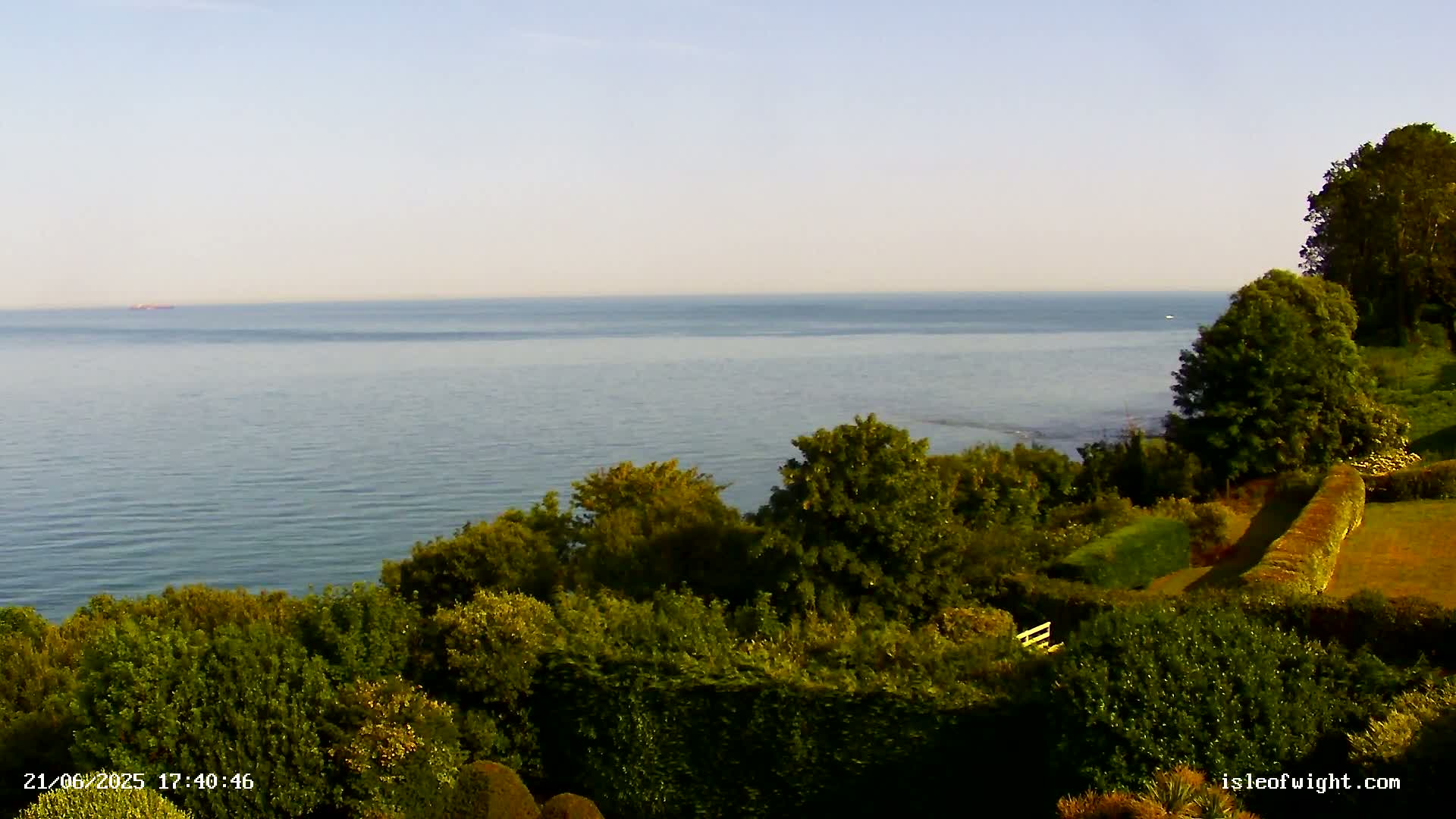 A calm sea is viewed from a verdant clifftop, with lush green vegetation and neatly trimmed hedges in the foreground.