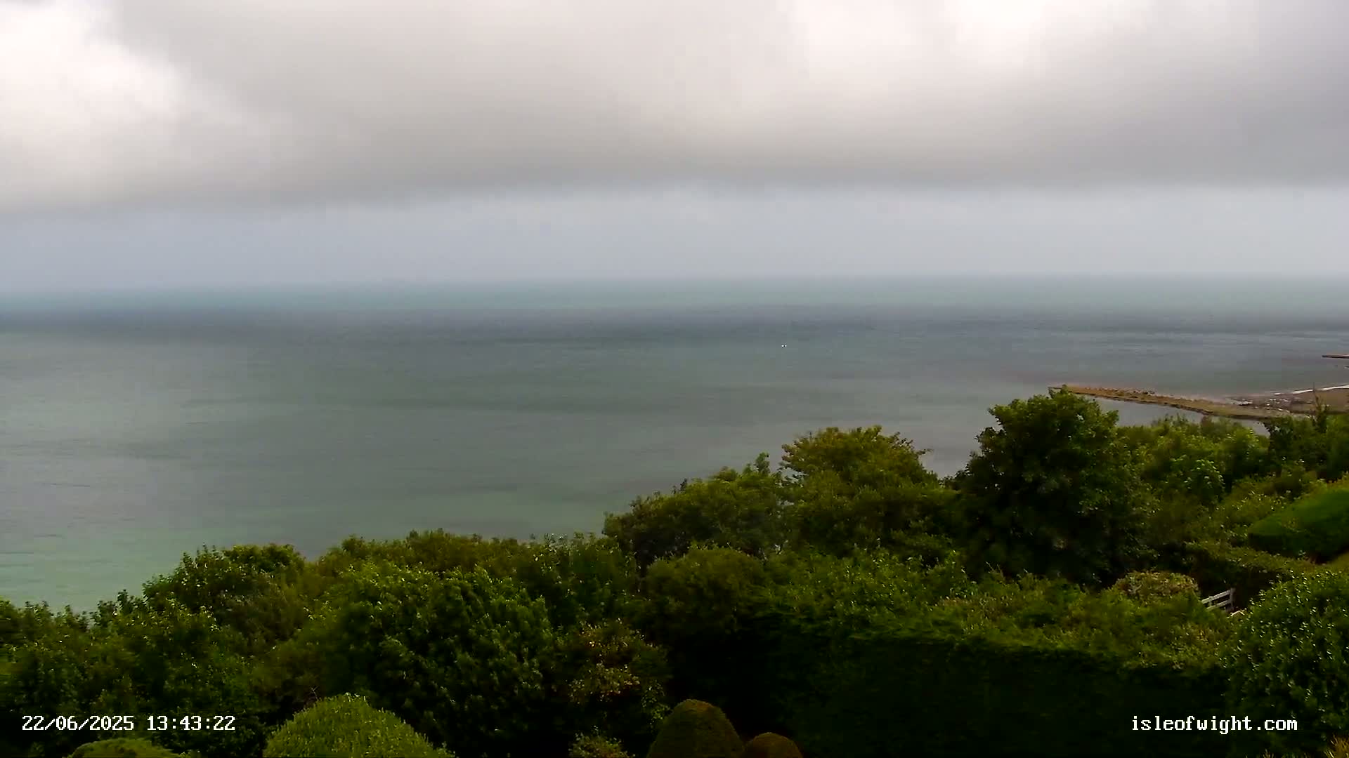 A calm, gray sea stretches to the horizon under an overcast sky, viewed from a high vantage point overlooking a lush green hillside.