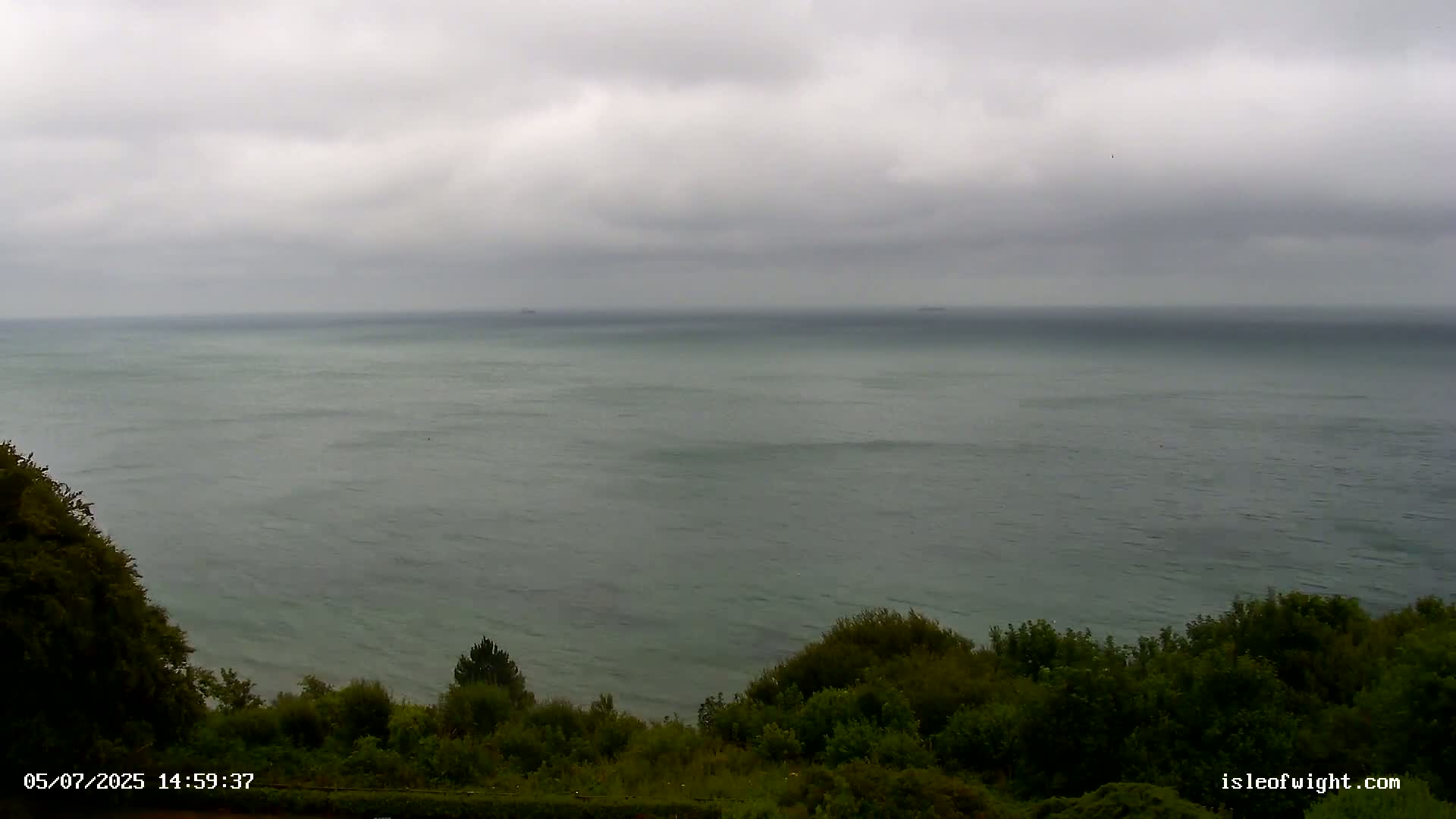 A calm, gray sea stretches to the horizon under an overcast sky, viewed from a vantage point above a line of green shrubbery.
