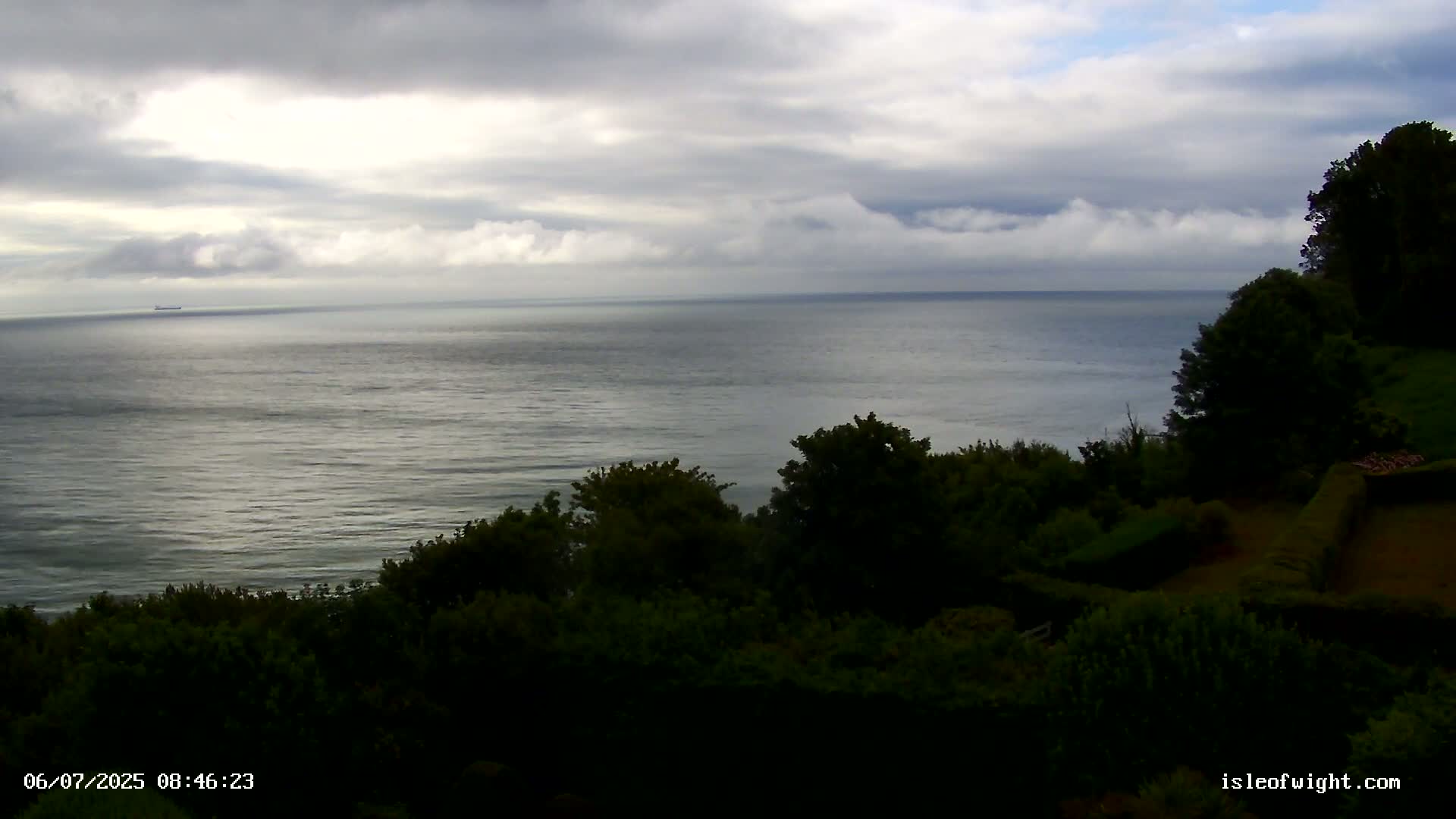A calm ocean stretches to the horizon under a cloudy sky, viewed from a vantage point above a dark, verdant hillside.