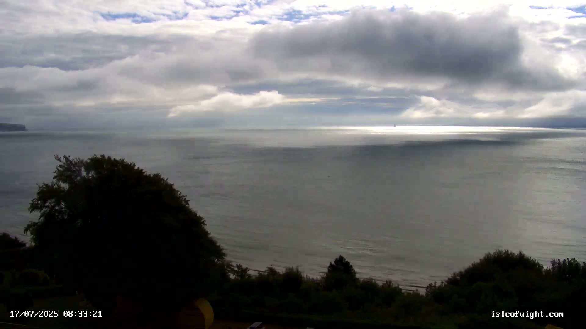 A mostly cloudy sky overlooks a calm, gray sea meeting a dark green shoreline with vegetation.