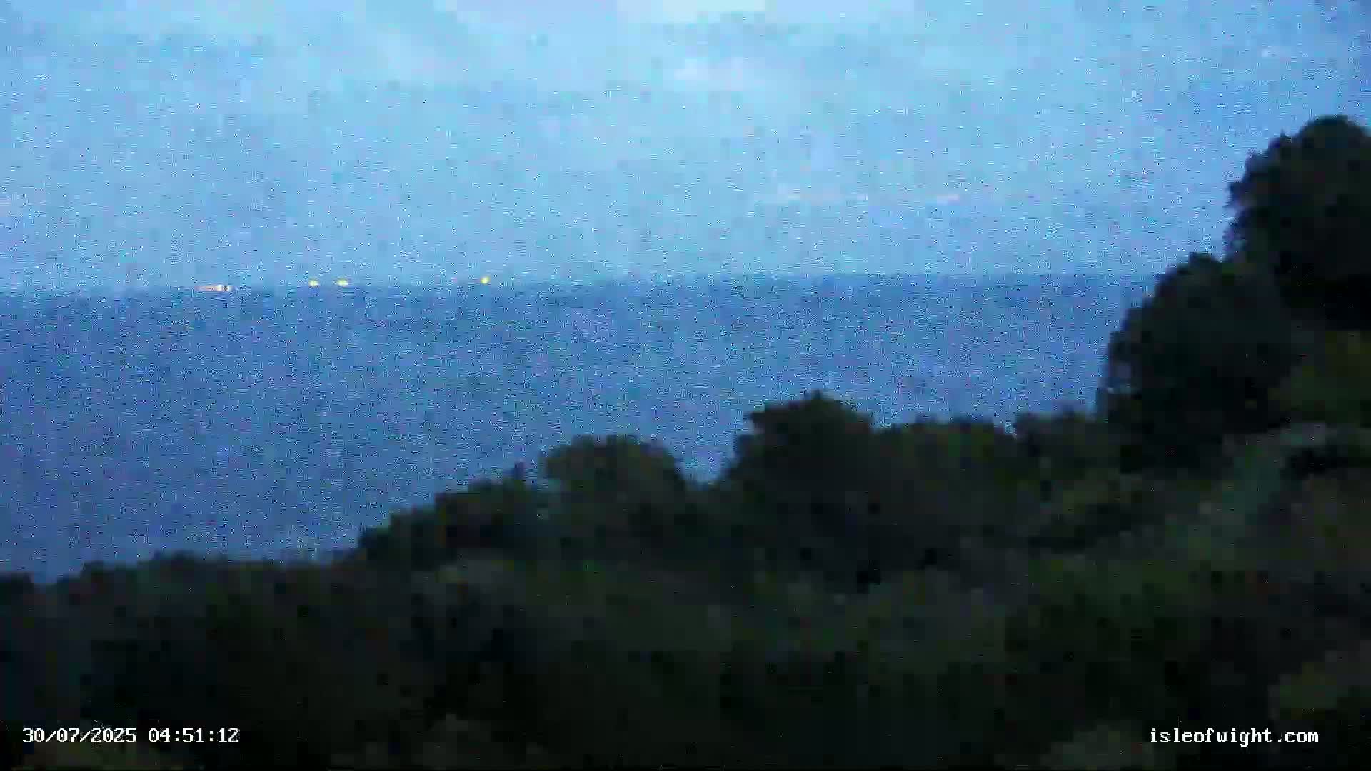A mostly cloudy sky overlooks a calm, gray sea meeting a dark green shoreline with vegetation.
