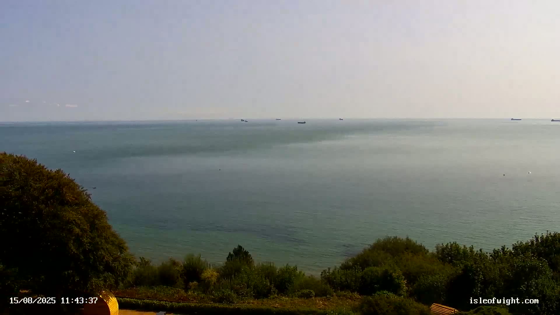 A mostly cloudy sky overlooks a calm, gray sea meeting a dark green shoreline with vegetation.