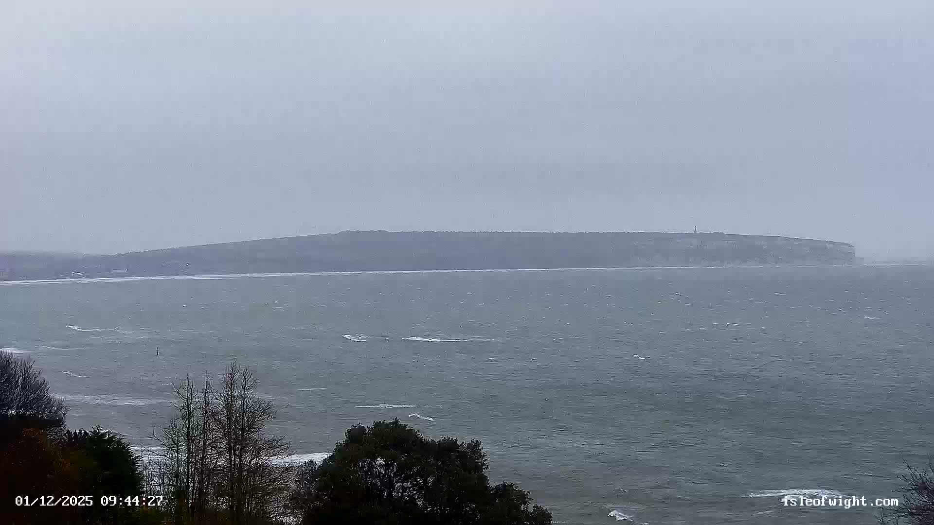 A wide shot captures a turbulent gray sea with numerous whitecaps under a heavily overcast sky, with a distant landmass featuring cliffs and a faint lighthouse, and a few foreground trees, indicating blustery and cold weather.