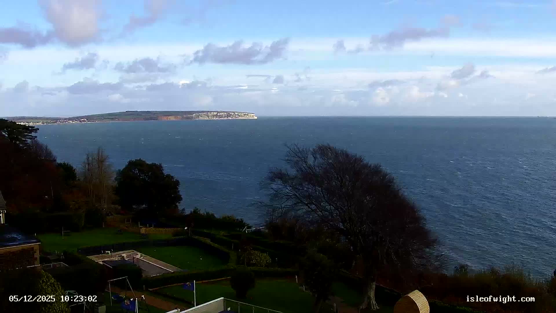 A scenic view on a bright, partly cloudy and breezy day overlooks a vast, choppy blue sea with a distant coastline featuring white cliffs and a coastal town, from a garden with manicured lawns and trees.