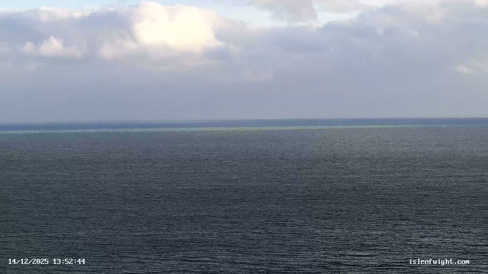 A scenic view on a bright, partly cloudy and breezy day overlooks a vast, choppy blue sea with a distant coastline featuring white cliffs and a coastal town, from a garden with manicured lawns and trees.