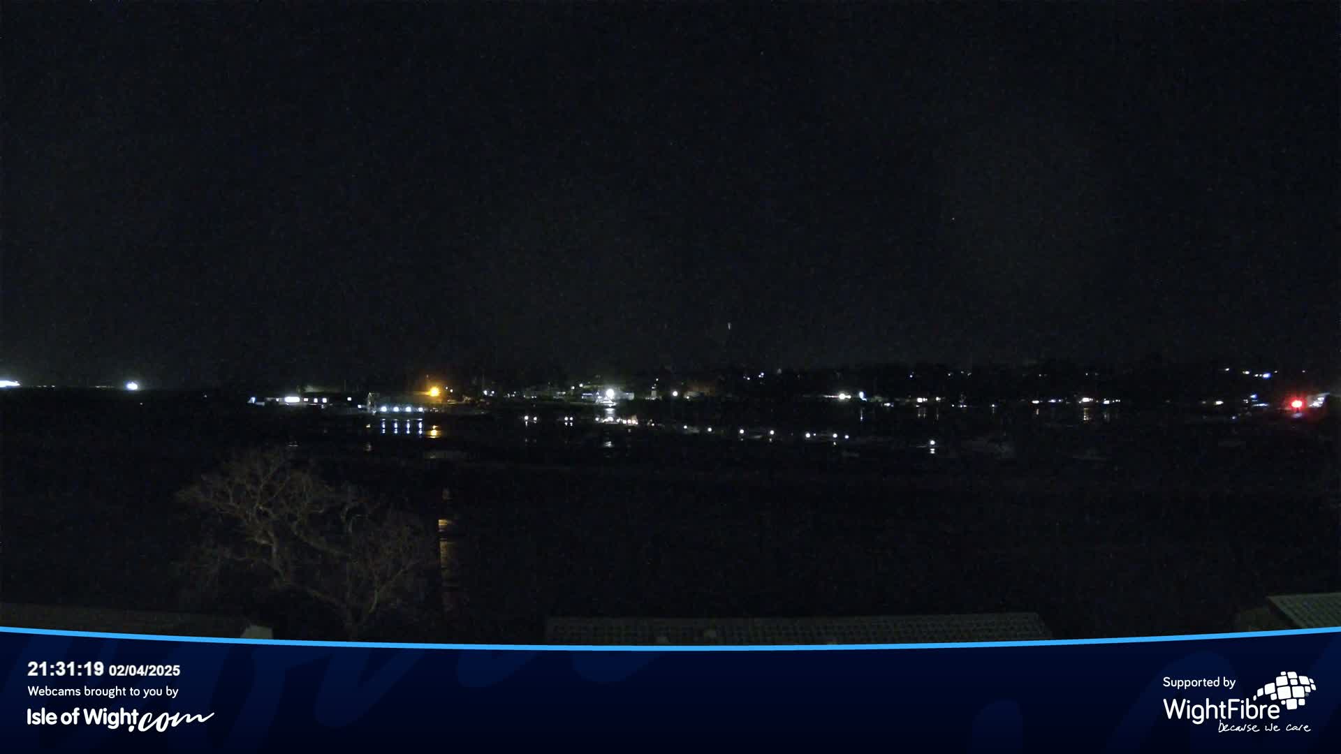 A nighttime view of a calm body of water reflecting the lights of a distant town under a clear, dark sky.