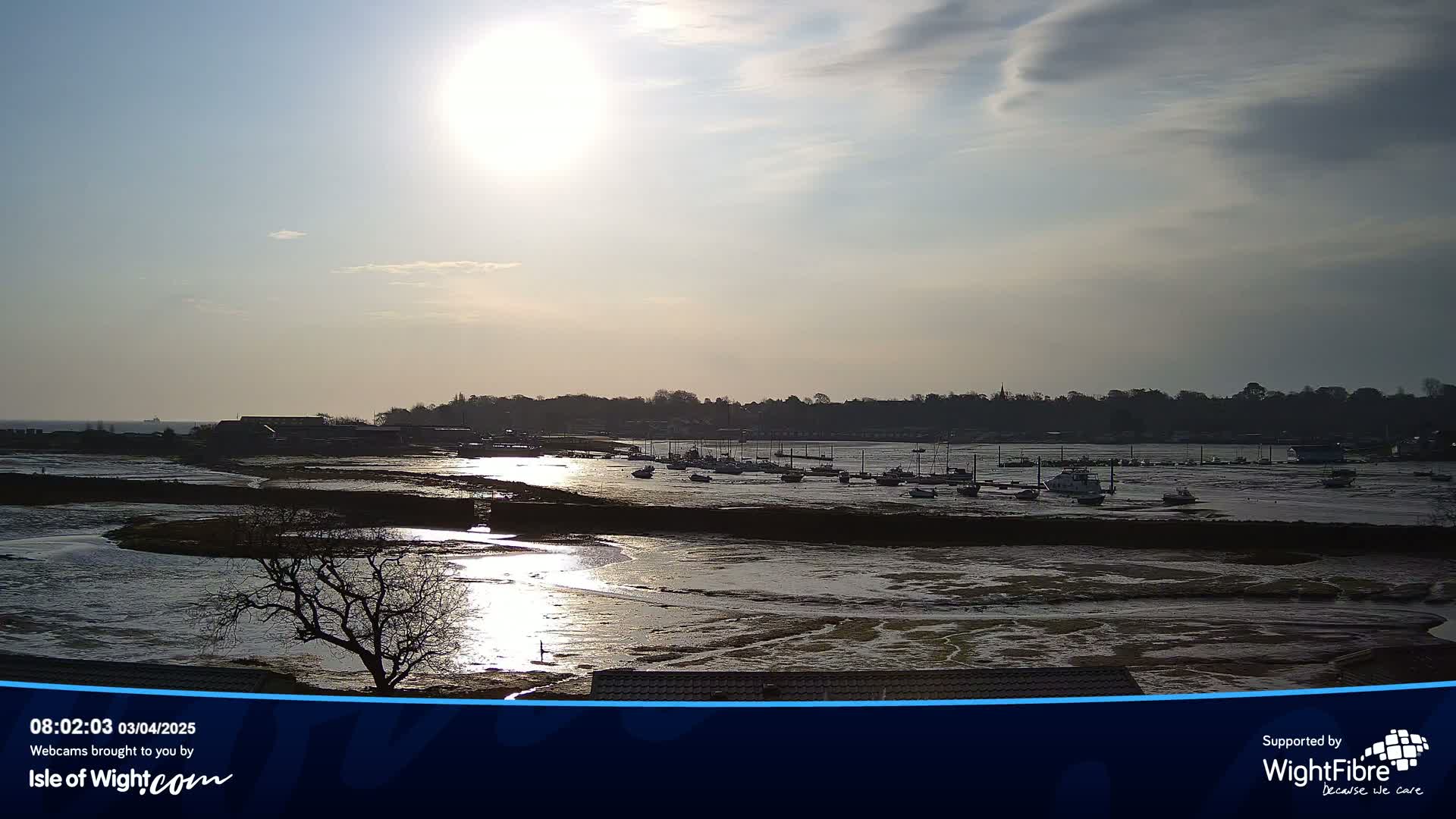 A partly sunny day reveals a wide tidal estuary with numerous boats moored at low tide, under a mostly cloudy sky.