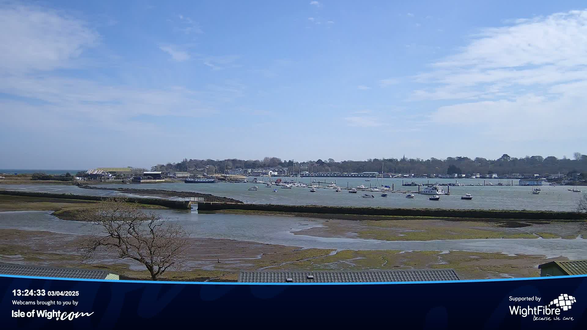A calm, sunny day reveals a harbor filled with boats, a marshy inlet, and a tree-lined shore in the distance.