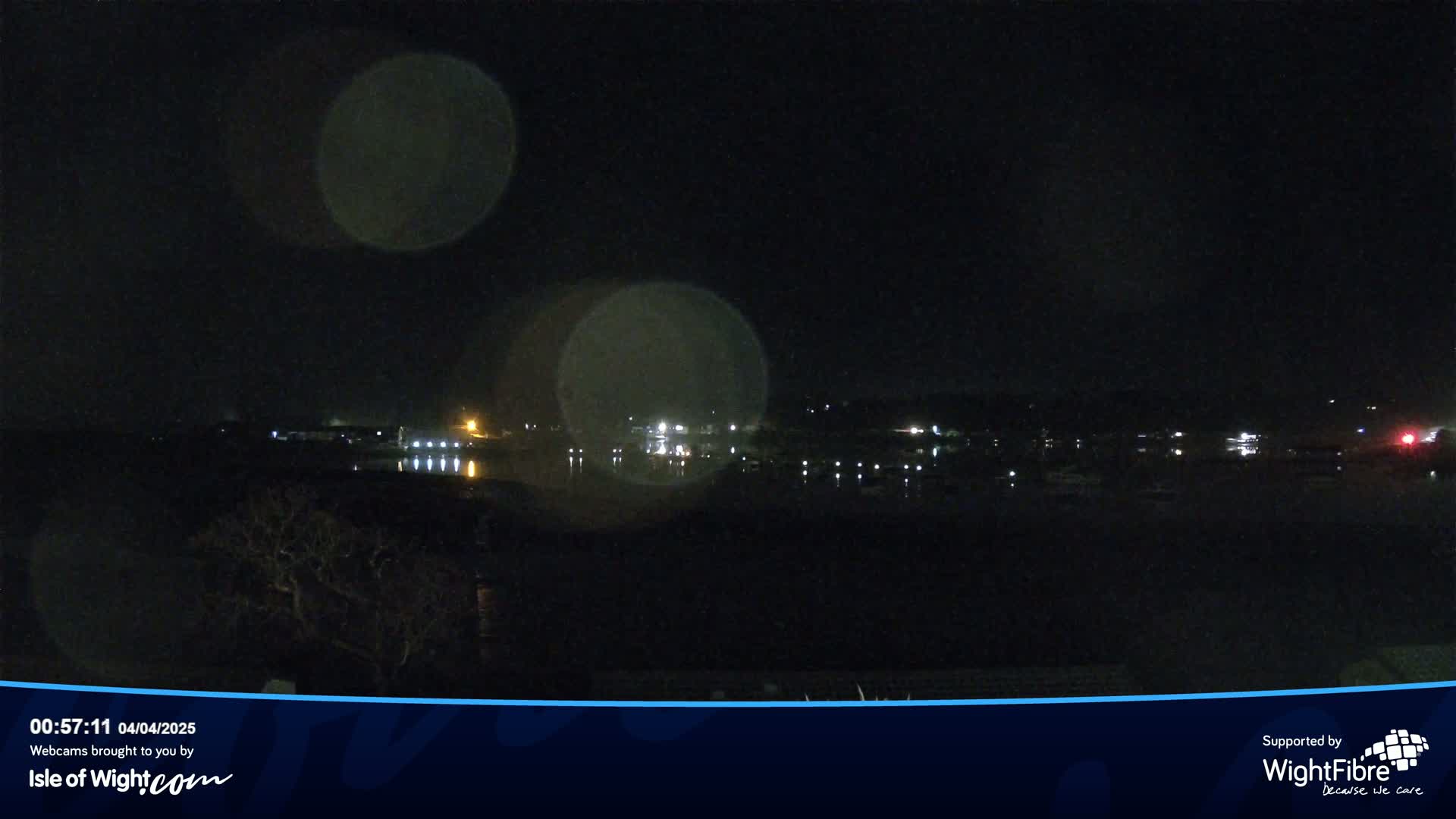 A nighttime view of a calm body of water with lights from a town visible across the water, under a dark, possibly rainy sky.