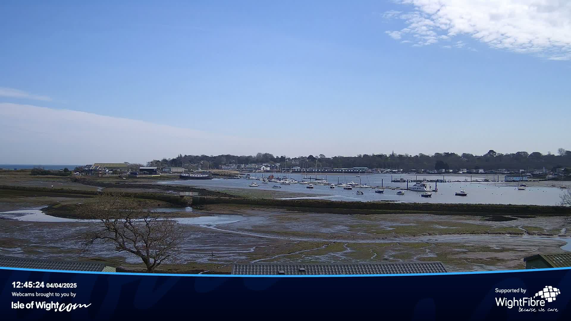 A mostly sunny day reveals a harbor filled with boats at low tide, with a village and trees visible in the background.