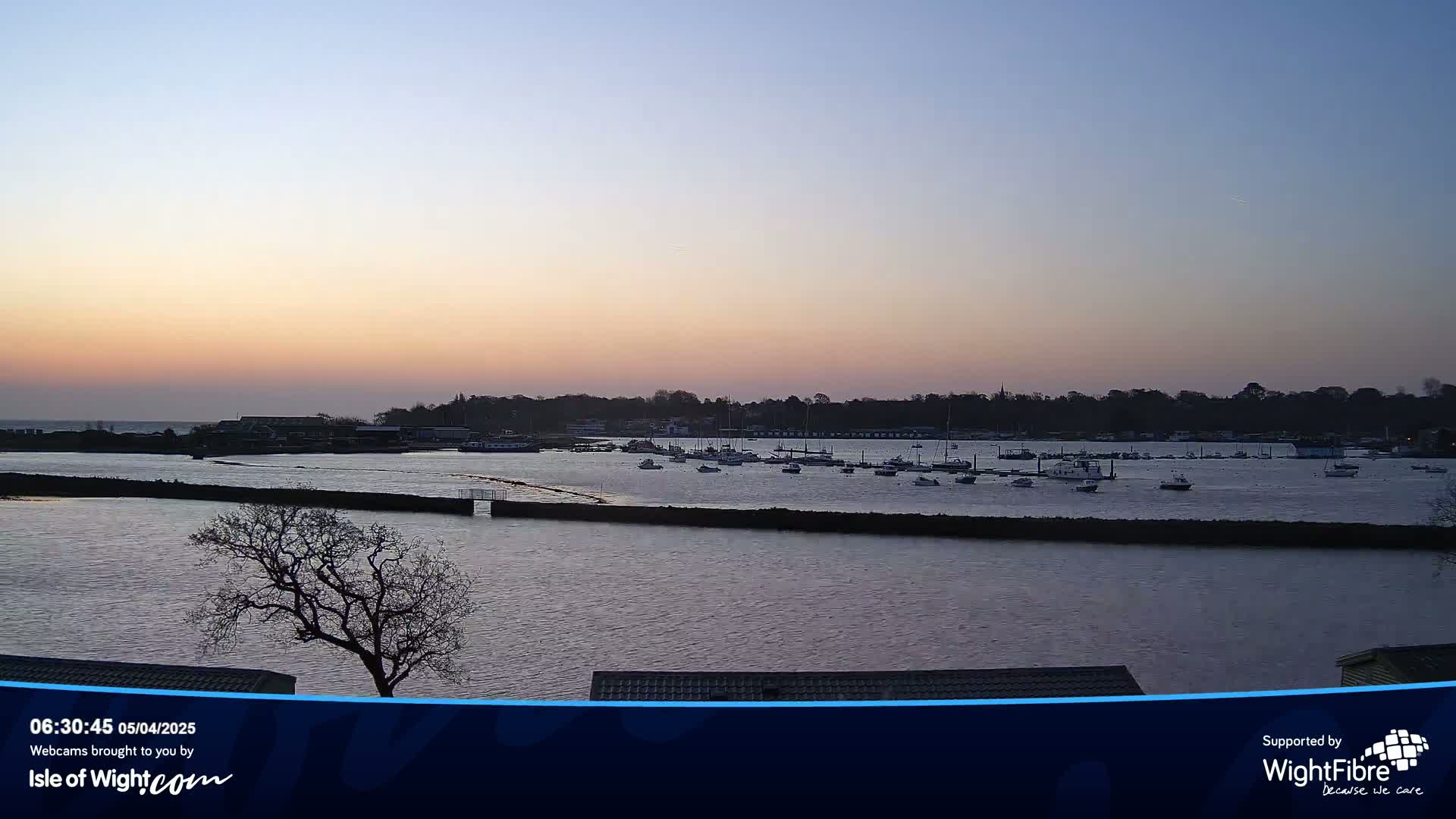 A calm body of water at sunset shows numerous boats moored near a shoreline with trees and buildings.