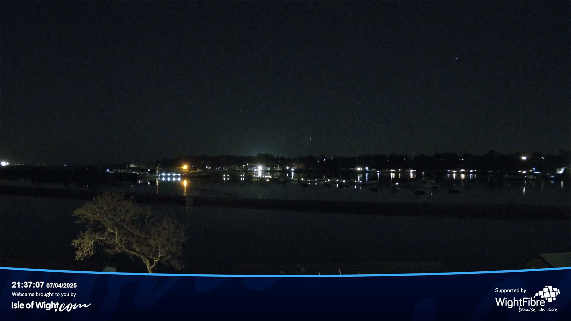 A nighttime view of a calm body of water reflecting the lights of a town across the shore, under a clear, dark sky.