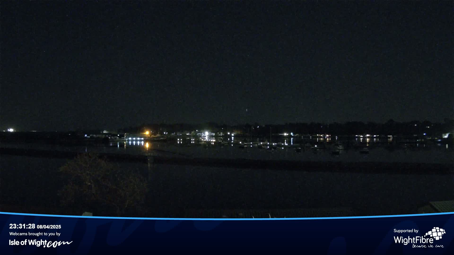 A nighttime view of a calm body of water with a line of lights reflecting on the surface, showing boats docked at a shoreline beyond.