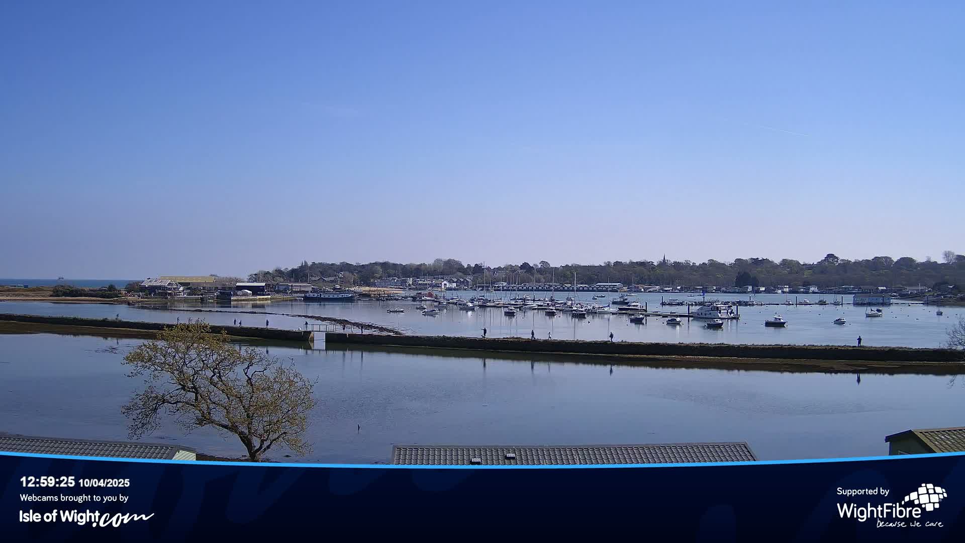 A calm, sunny day reveals a harbor filled with boats, a shoreline with buildings and trees, and a tranquil waterway leading to the ocean.