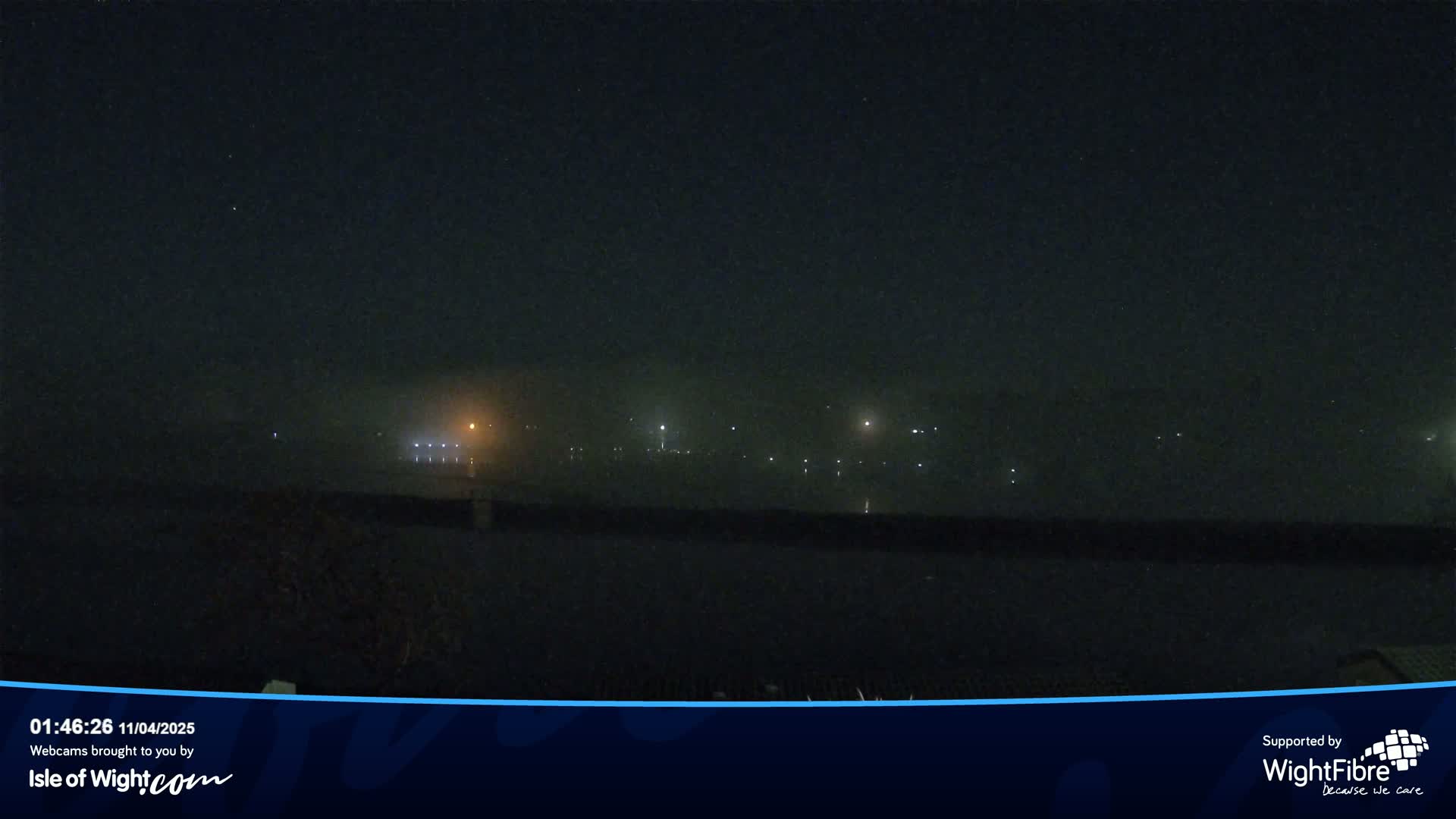 A foggy nighttime scene shows a body of water with faint lights visible across it in the distance.
