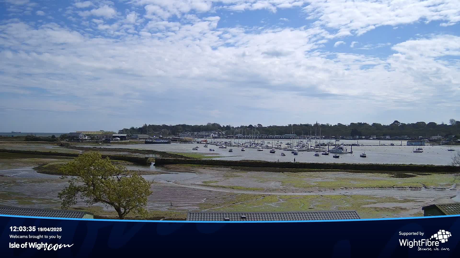 A mostly sunny day reveals a harbor filled with numerous boats at low tide, with a small town visible in the background.
