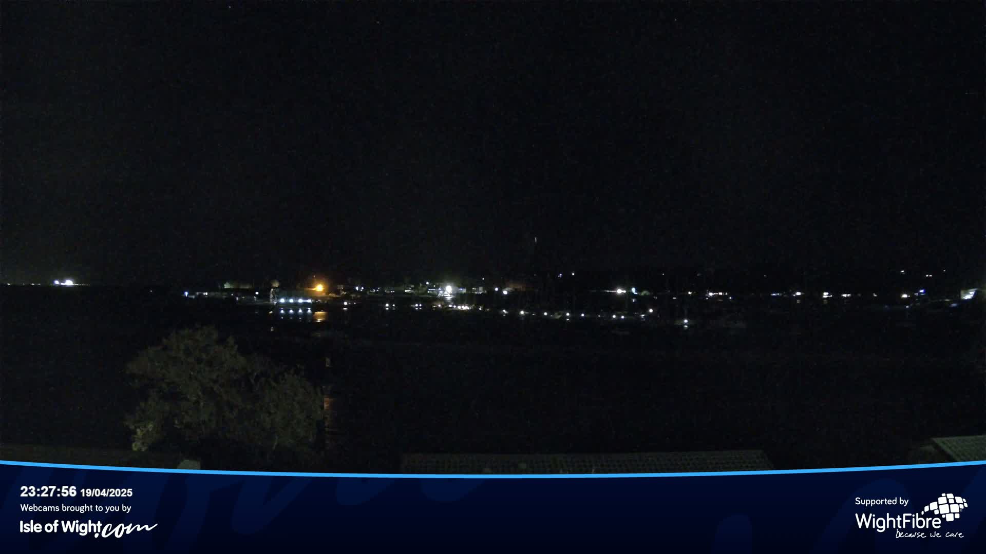 A dark nighttime scene shows a body of water with a line of dimly lit buildings and trees across it under a clear, starless sky.