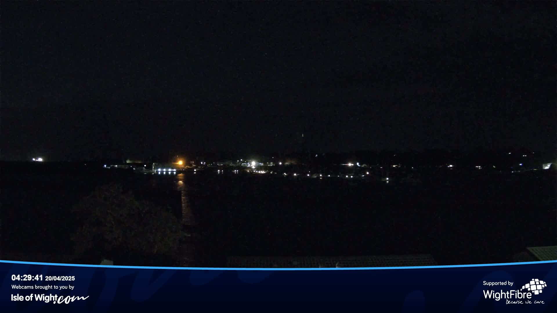 A nighttime view of a dark body of water with faintly lit buildings and boats on the opposite shore under a clear, dark sky.