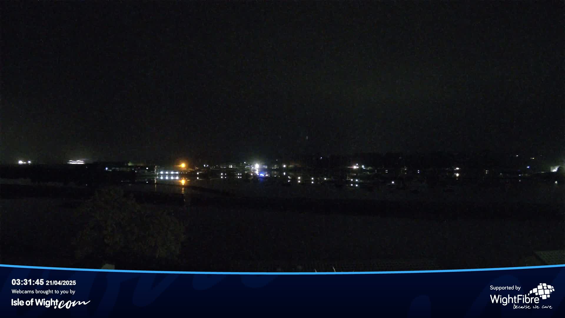 A nighttime long shot of a dark harbor with dimly lit boats and a town in the background under a clear, dark sky.