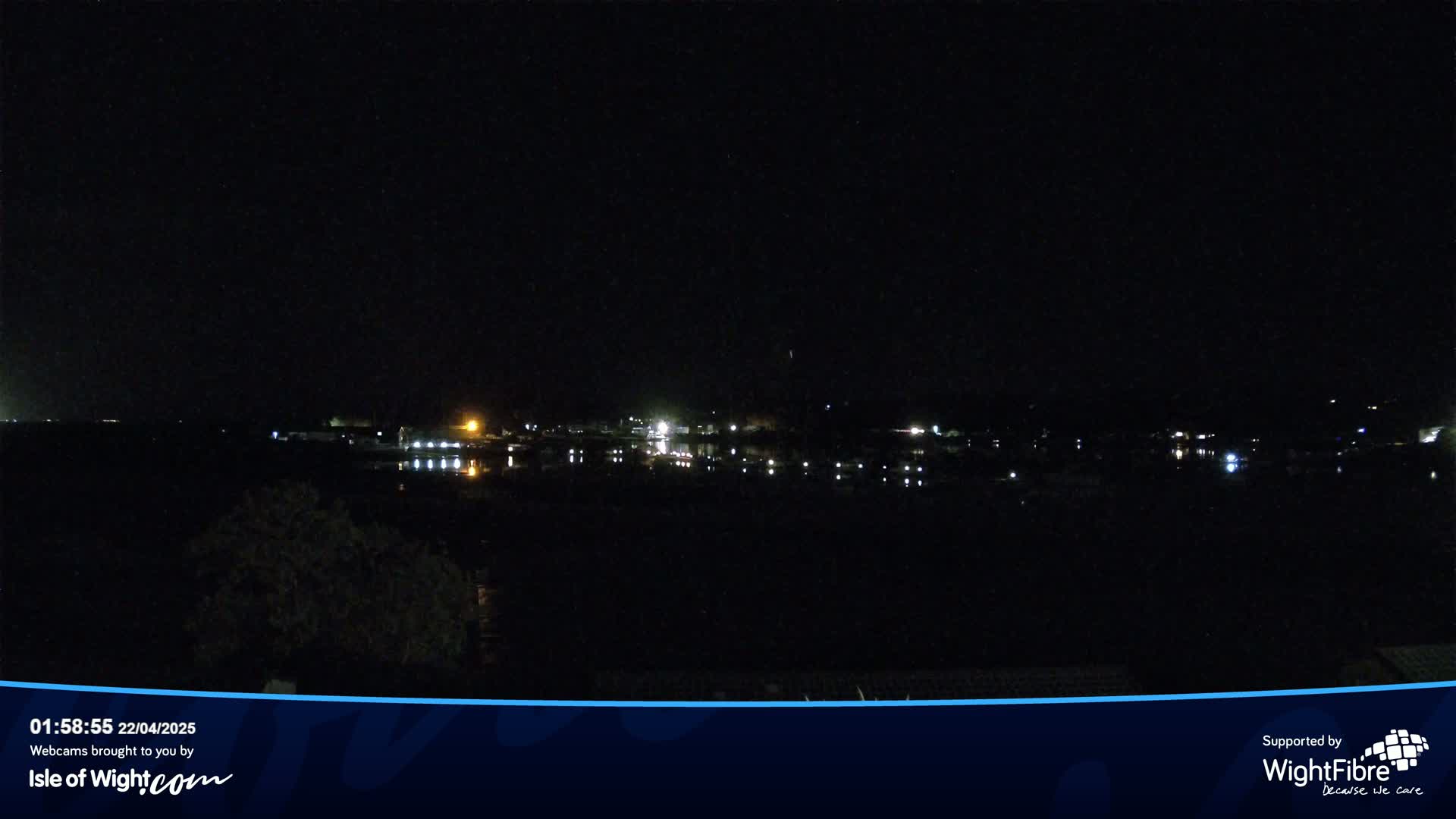 A nighttime view of a calm body of water reflecting the lights of a town across the water under a dark, clear sky.