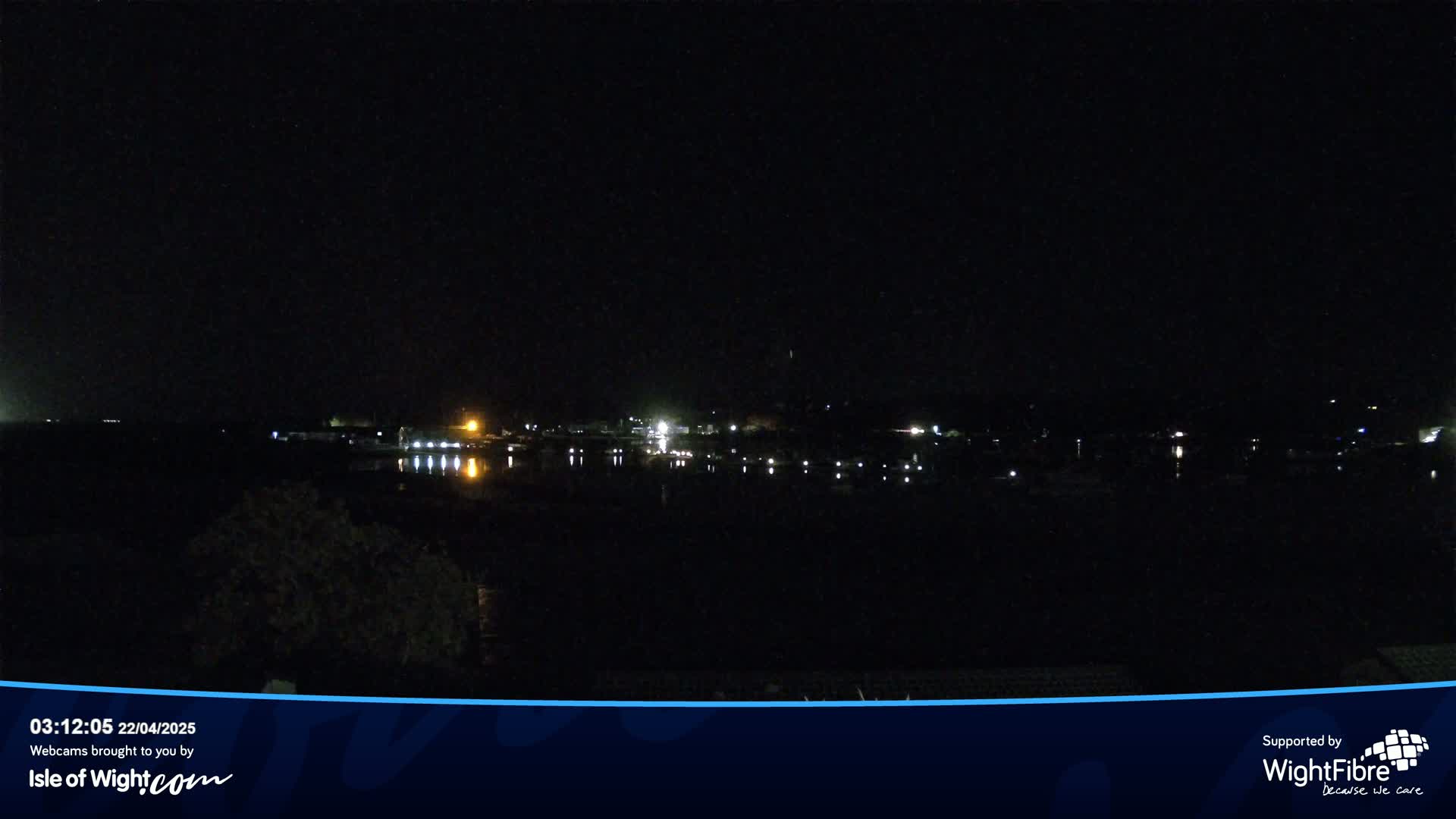 A nighttime long shot of a dark body of water reflecting the lights of a distant town under a clear, black sky.