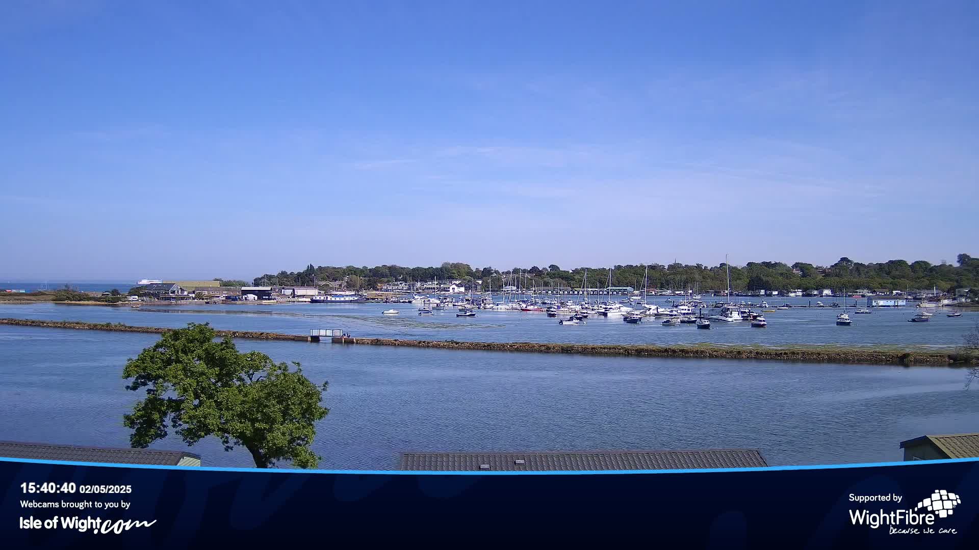 A calm, sunny day reveals a marina filled with numerous boats, situated across a waterway from a verdant shoreline.