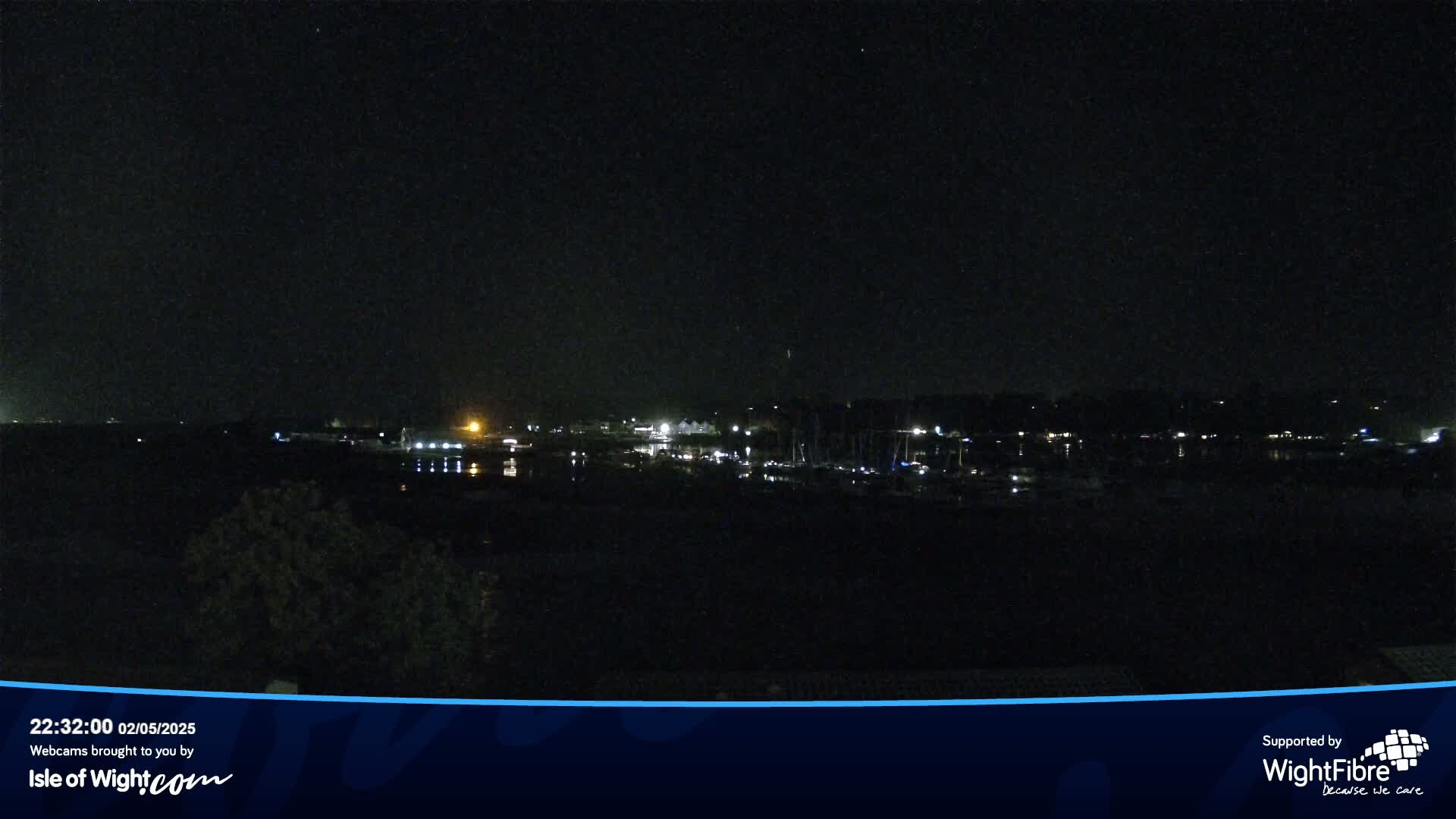 A nighttime view of a calm body of water with a line of lights from buildings and boats across the far shore under a clear, dark sky.