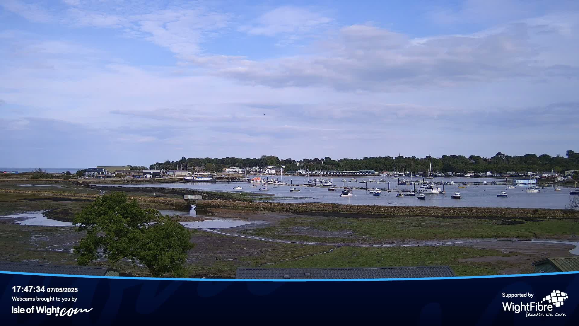 Under a partly cloudy sky, a calm body of water shows many boats moored in a harbor near a shoreline with low-lying vegetation.
