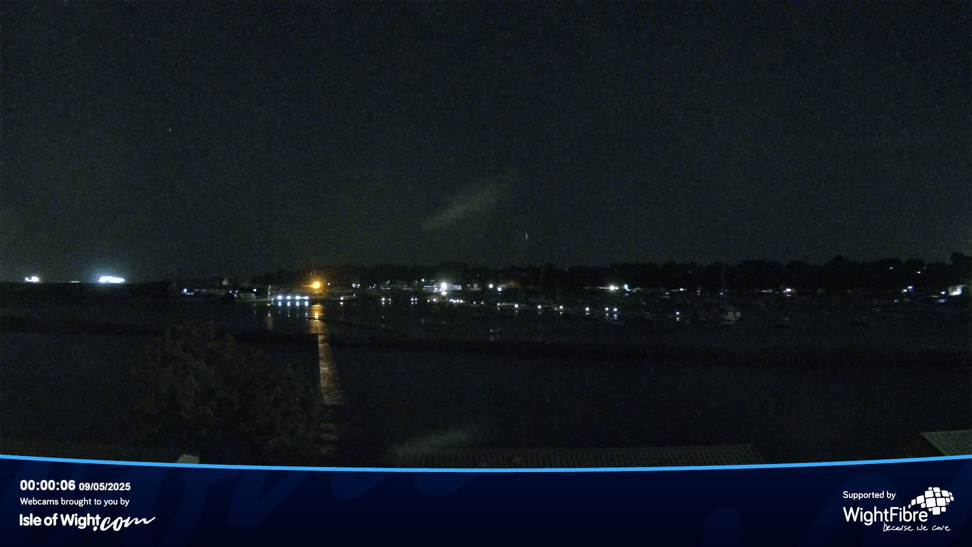 A nighttime view of a calm harbor with docked boats and dimly lit buildings across the water under a clear, dark sky.