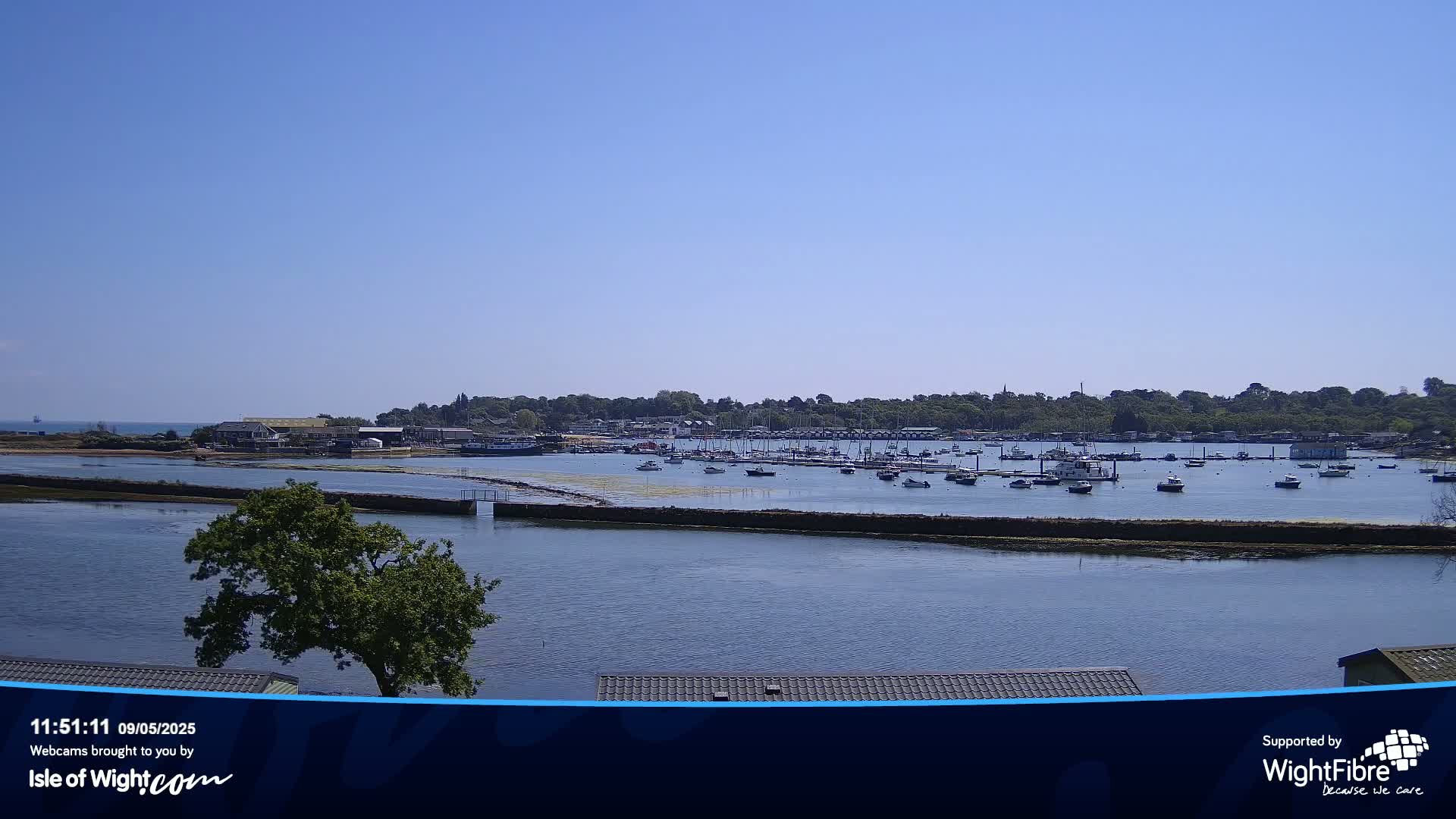 A calm, sunny day reveals a harbor filled with numerous boats, situated near a tree-lined shoreline.