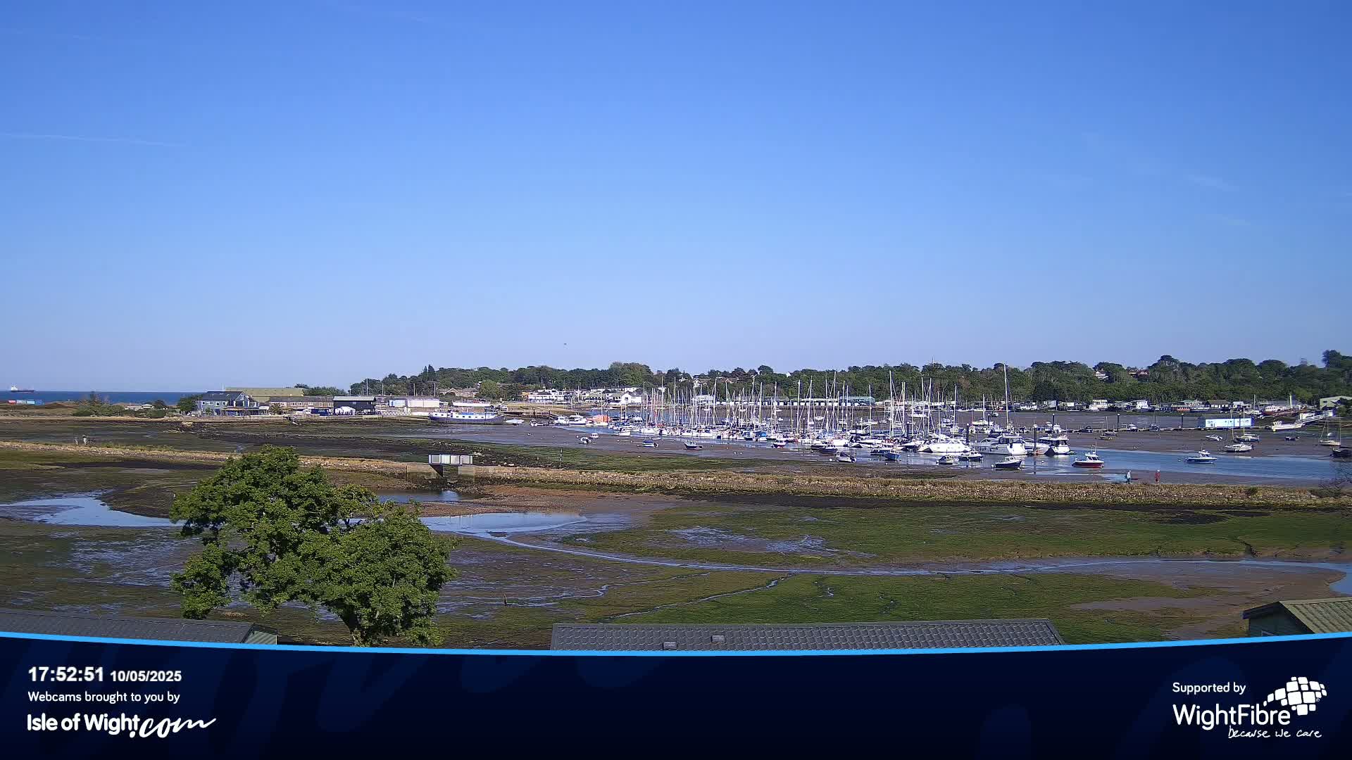 Under a clear blue sky, a marina filled with numerous sailboats is situated next to a vast expanse of exposed mudflats and shallow water.