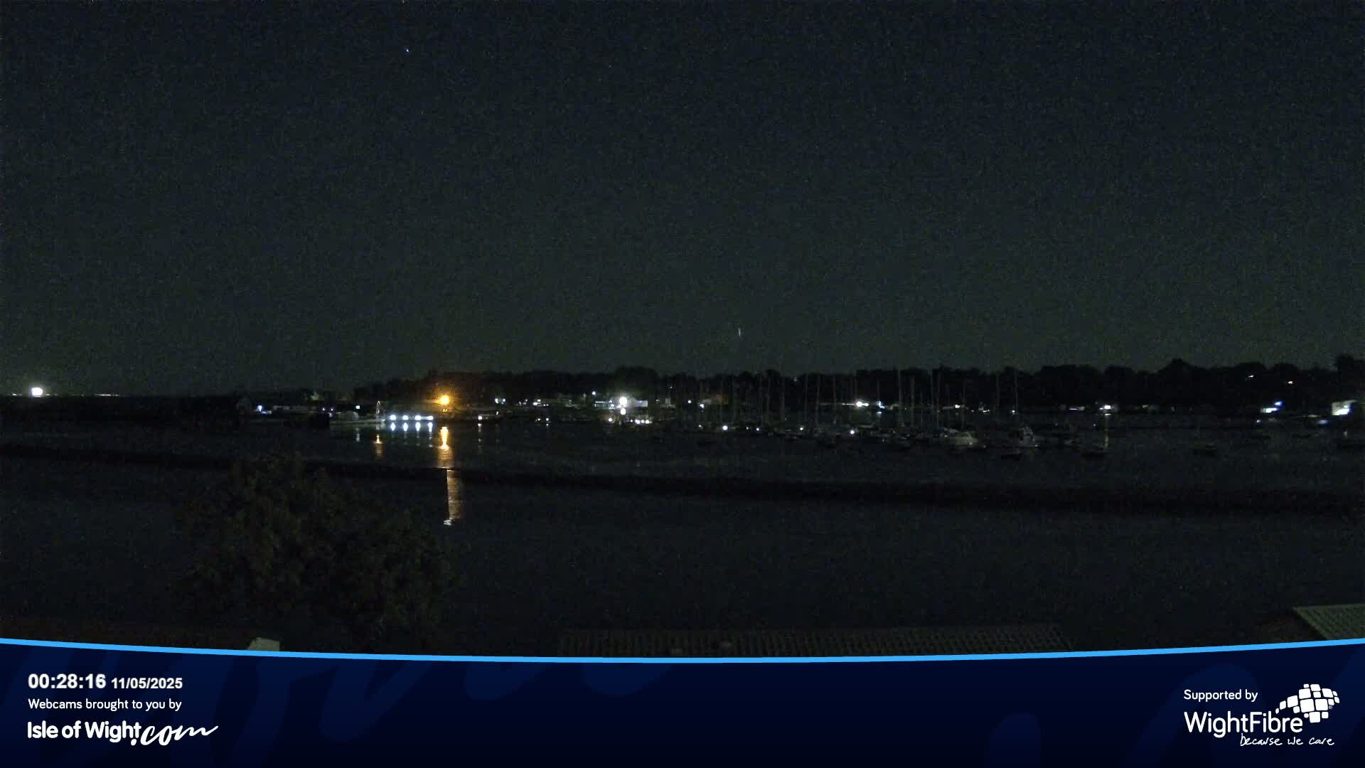 A nighttime view of a calm body of water with docked boats and a dark tree line in the background under a clear, starlit sky.