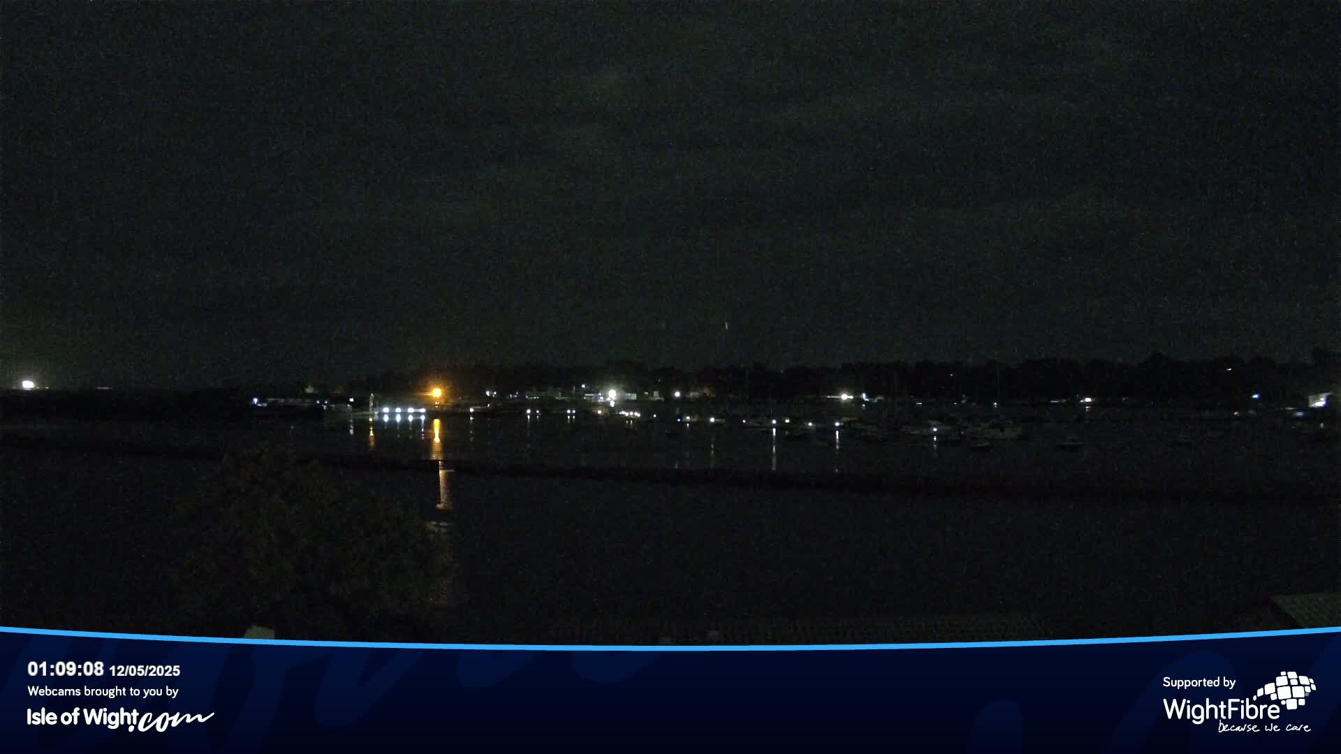 A nighttime view of a calm body of water with boats moored along a shoreline, illuminated by faint lights from buildings and boats.