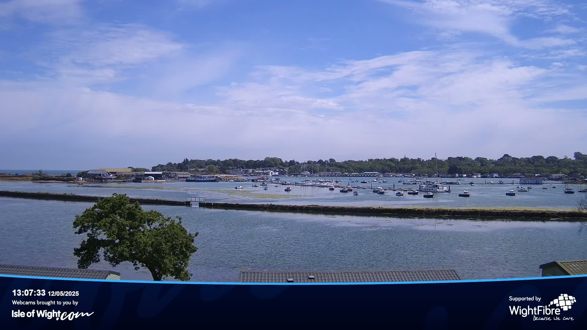 A calm body of water with numerous boats in a marina, under a partly cloudy sky, is bordered by green land.