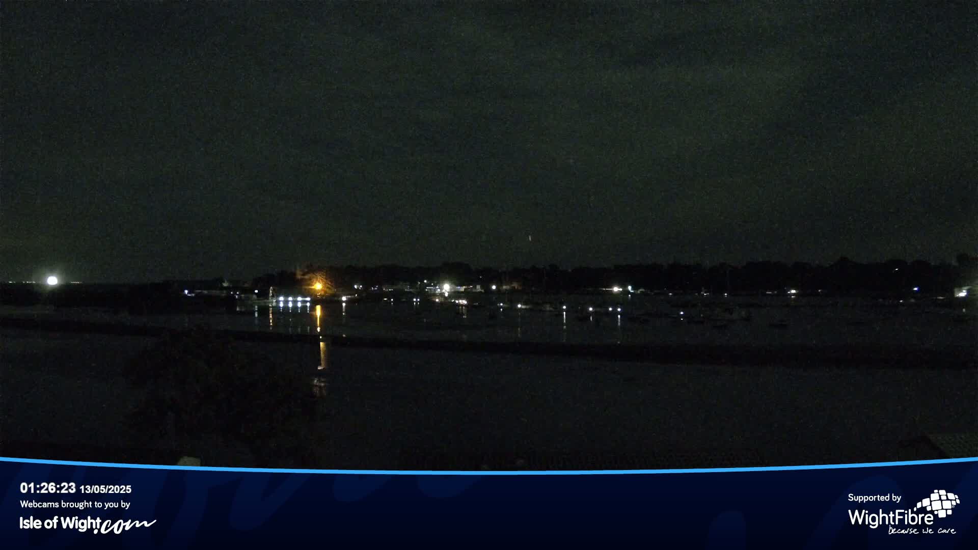 A dark nighttime scene shows a calm body of water reflecting the faint lights of a distant shoreline.